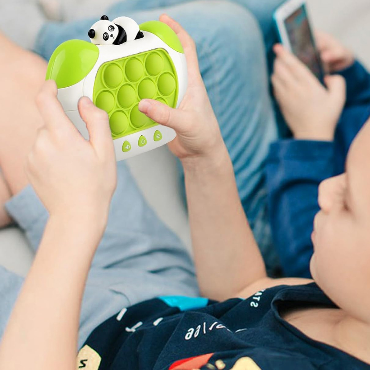 Un niño sosteniendo un juguete pop-it con forma de panda verde y blanco. Otra persona usa un teléfono.