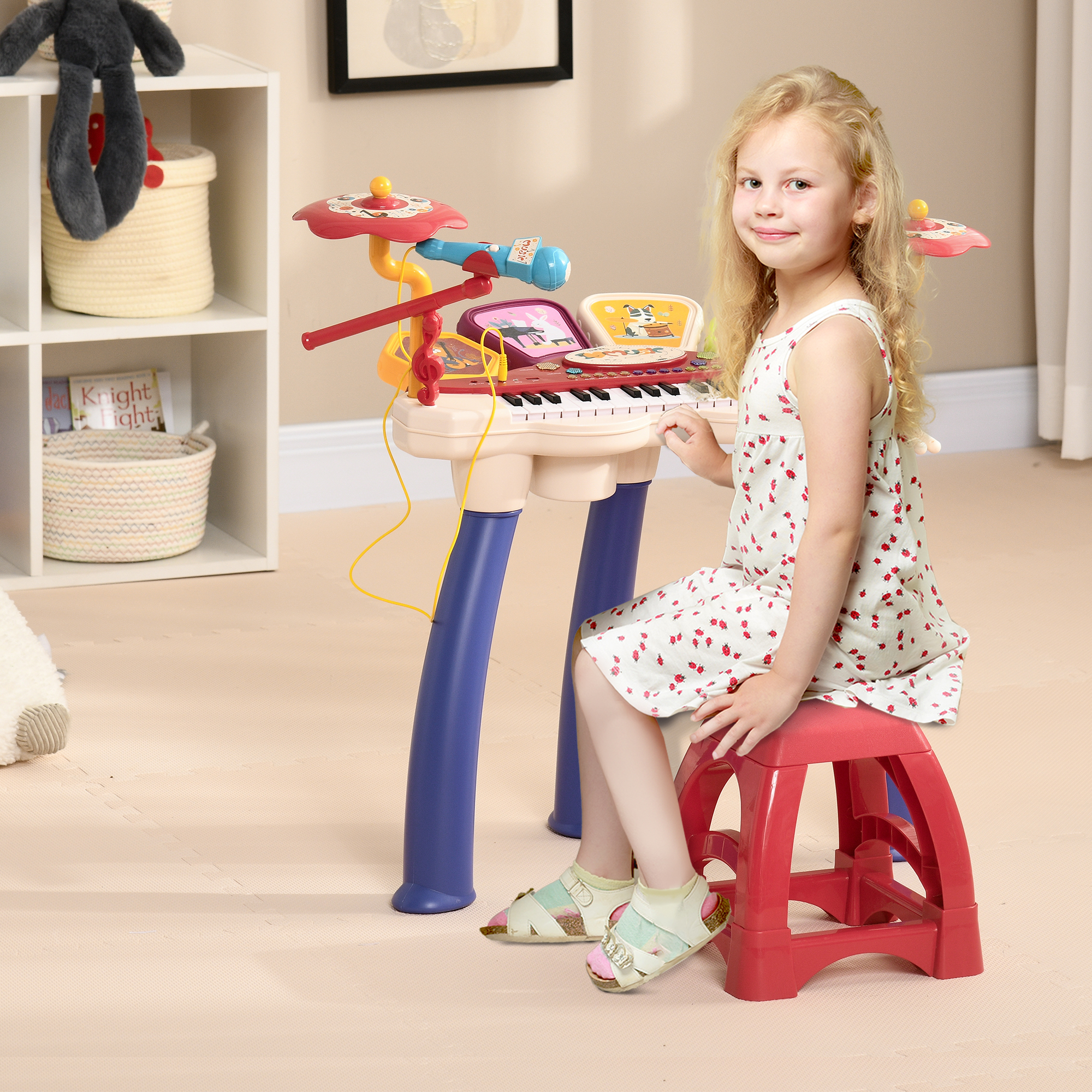 Una niña pequeña está sentada junto a un piano de juguete, sonriendo a la cámara. Hay un taburete y una batería de juguete.