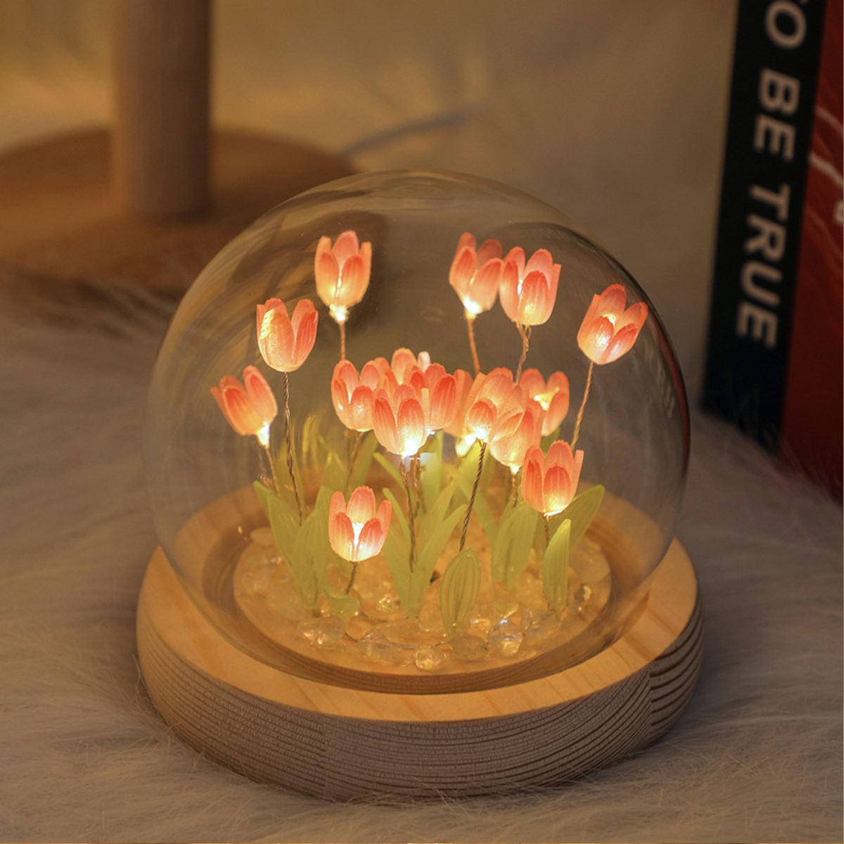Glaskuppel mit beleuchteten rosa Tulpen, auf Holzsockel, mit Buch im Hintergrund.
