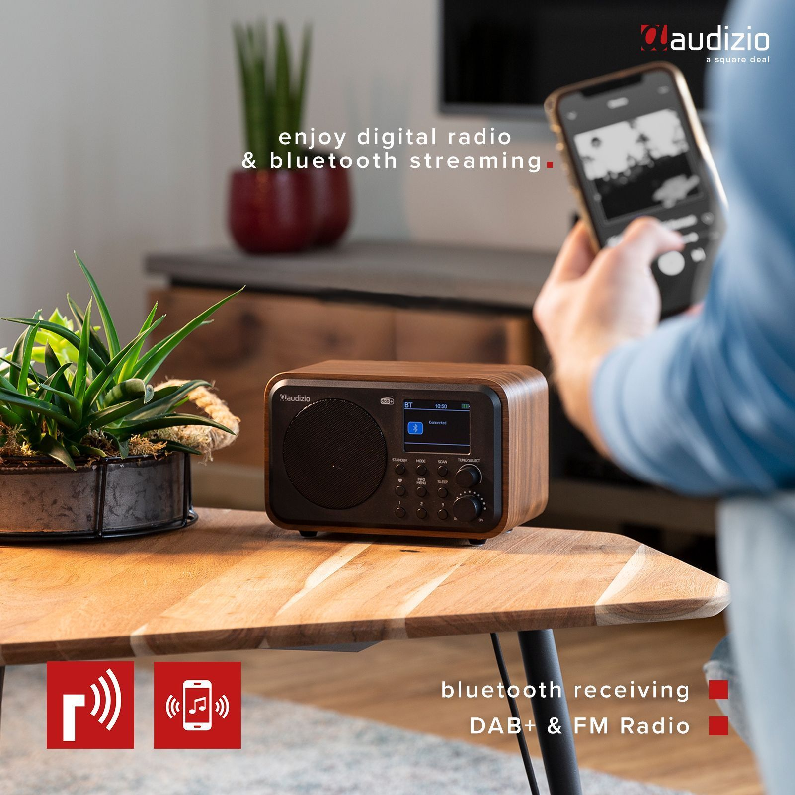 Ein Holzradio auf einem Holztisch, eine Person hält ein Telefon. Das Radio hat einen Bildschirm.