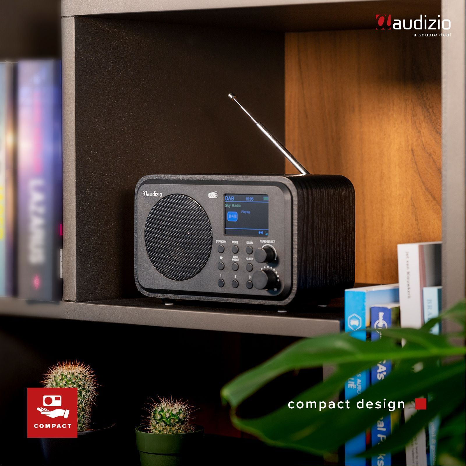 Una radio negra con pantalla en una estantería. Hay libros y cactus en el fondo. El texto dice 'diseño compacto'.