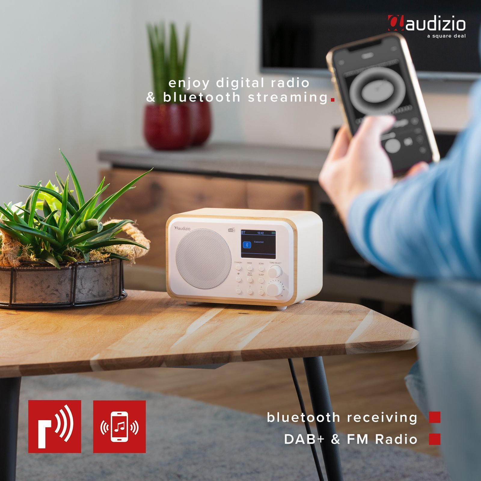 Radio blanca sobre una mesa de madera, una persona sosteniendo un smartphone al fondo.