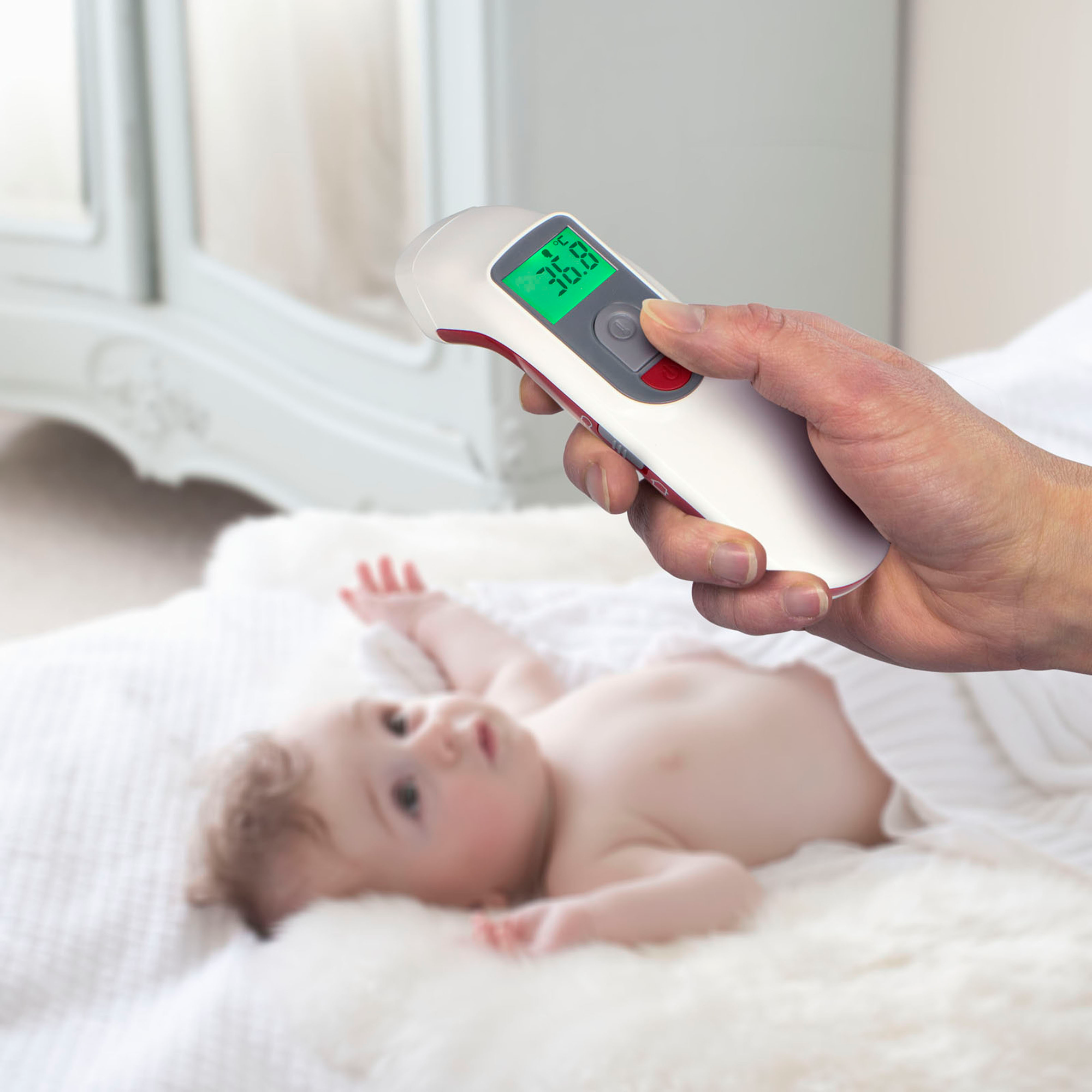 Die Hand einer Person, die ein Thermometer über einem Baby hält, das auf einem Bett liegt.