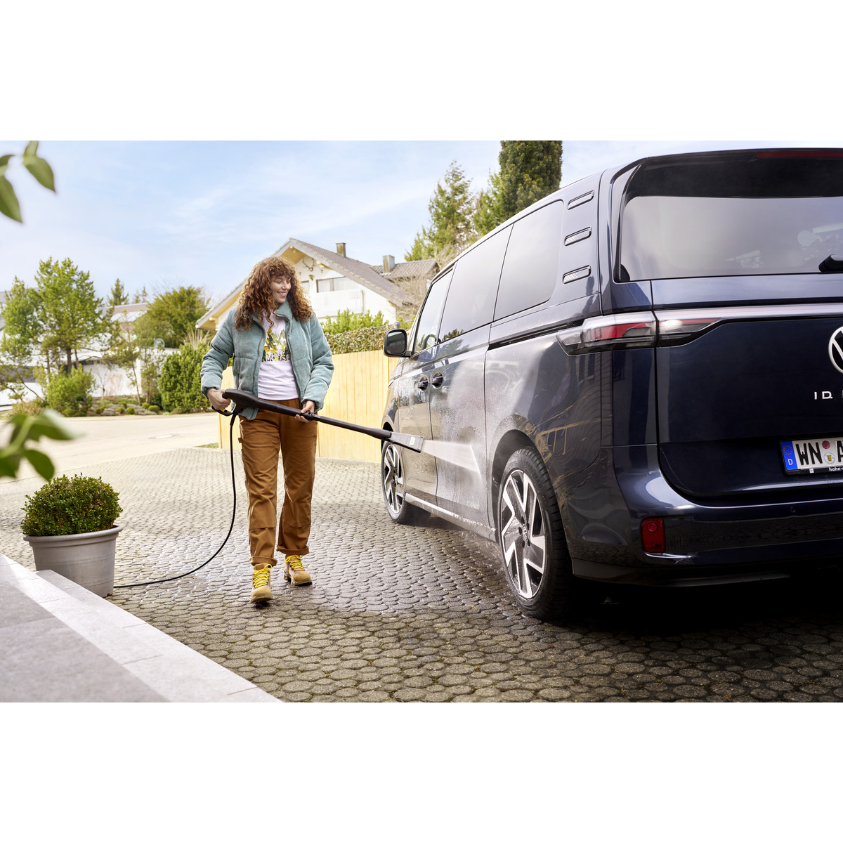 Kobieta myjąca niebieski samochód wężem, stojąca na kamiennym chodniku z zielonym krzewem.