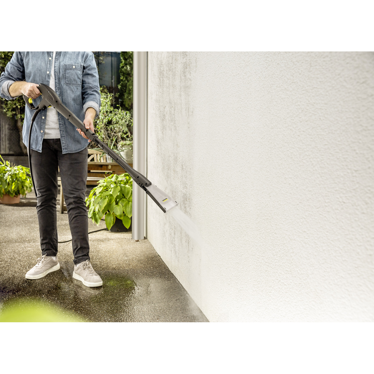Mężczyzna w dżinsach czyści białą ścianę myjką ciśnieniową. Zielone rośliny. Betonowa podłoga.