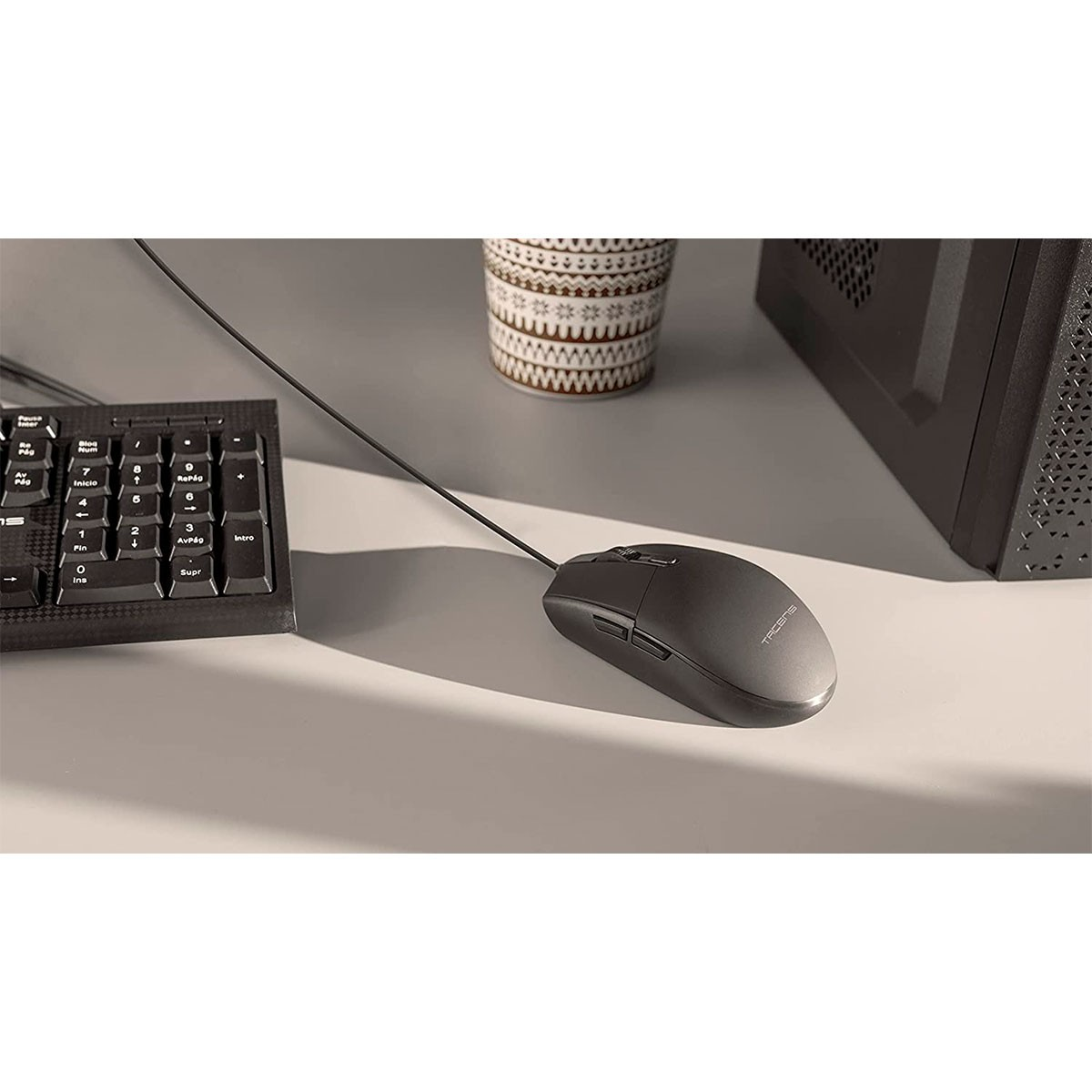 A computer mouse with a cable, on a white desk. A keyboard and a computer are visible in the background.