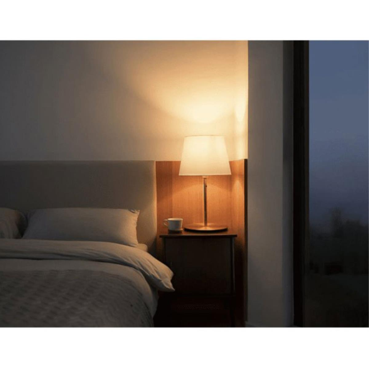 Una lampada da comodino accesa con un paralume bianco accanto a un letto in una stanza poco illuminata.