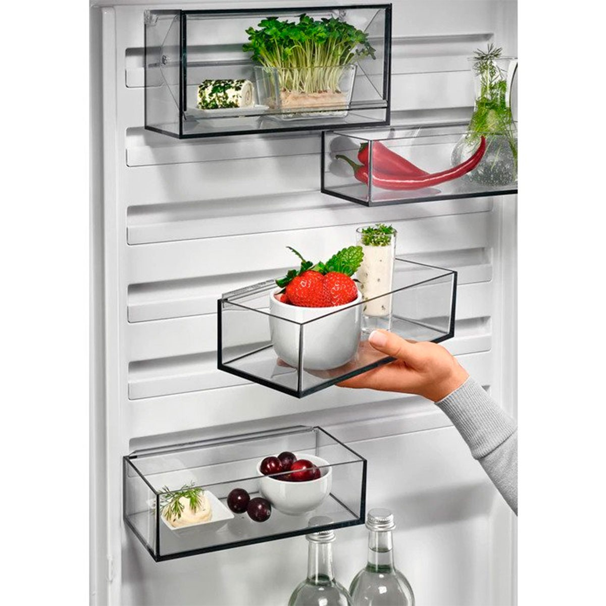 Interior de nevera con estantes transparentes que contienen alimentos. Una mano alcanza un estante.