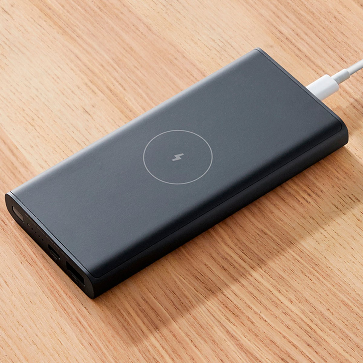 Un power bank grigio scuro su una superficie di legno, con un simbolo di ricarica e un cavo di ricarica.