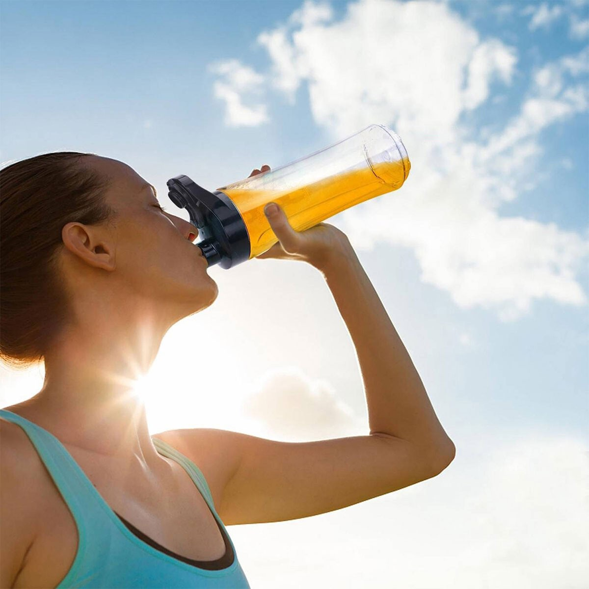 Eine Frau trinkt aus einer klaren Flasche mit orangefarbener Flüssigkeit unter sonnigem Himmel.