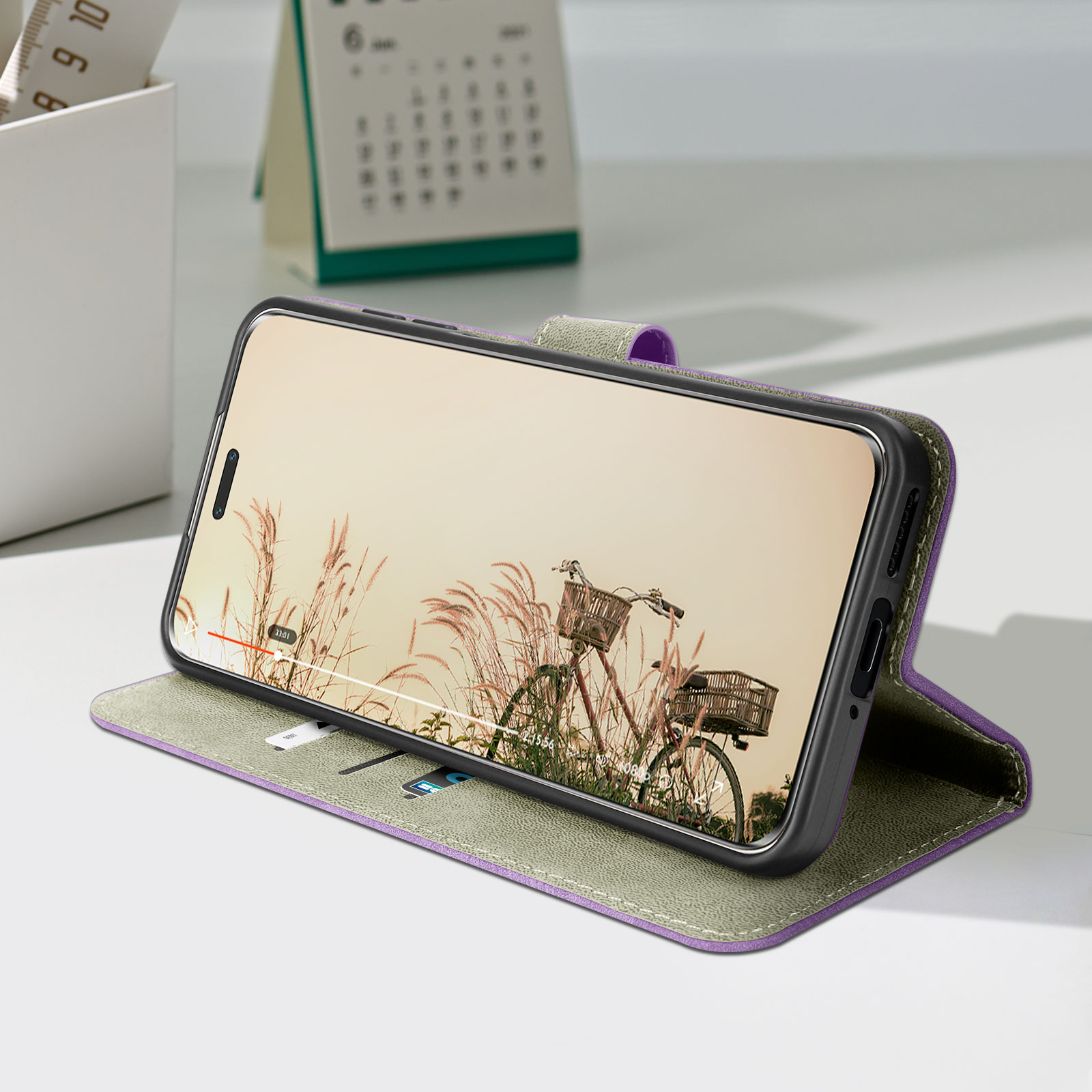 Telefono in custodia, aperto su un tavolo. Il display mostra una scena di biciclette. Calendario e righello sullo sfondo.
