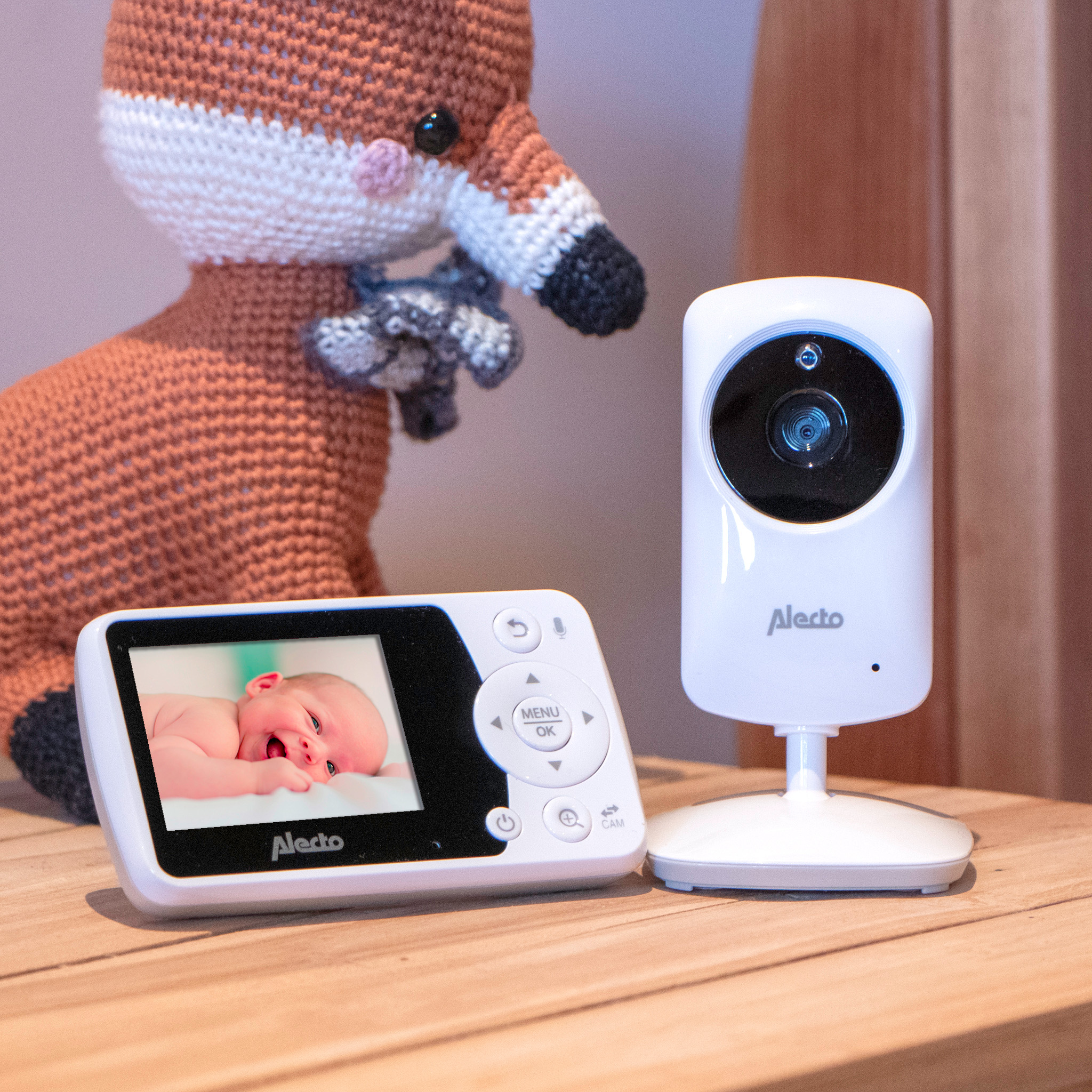 Een babyfoonset met een lachende baby op het scherm, een vossenspeeltje, op een houten tafel.