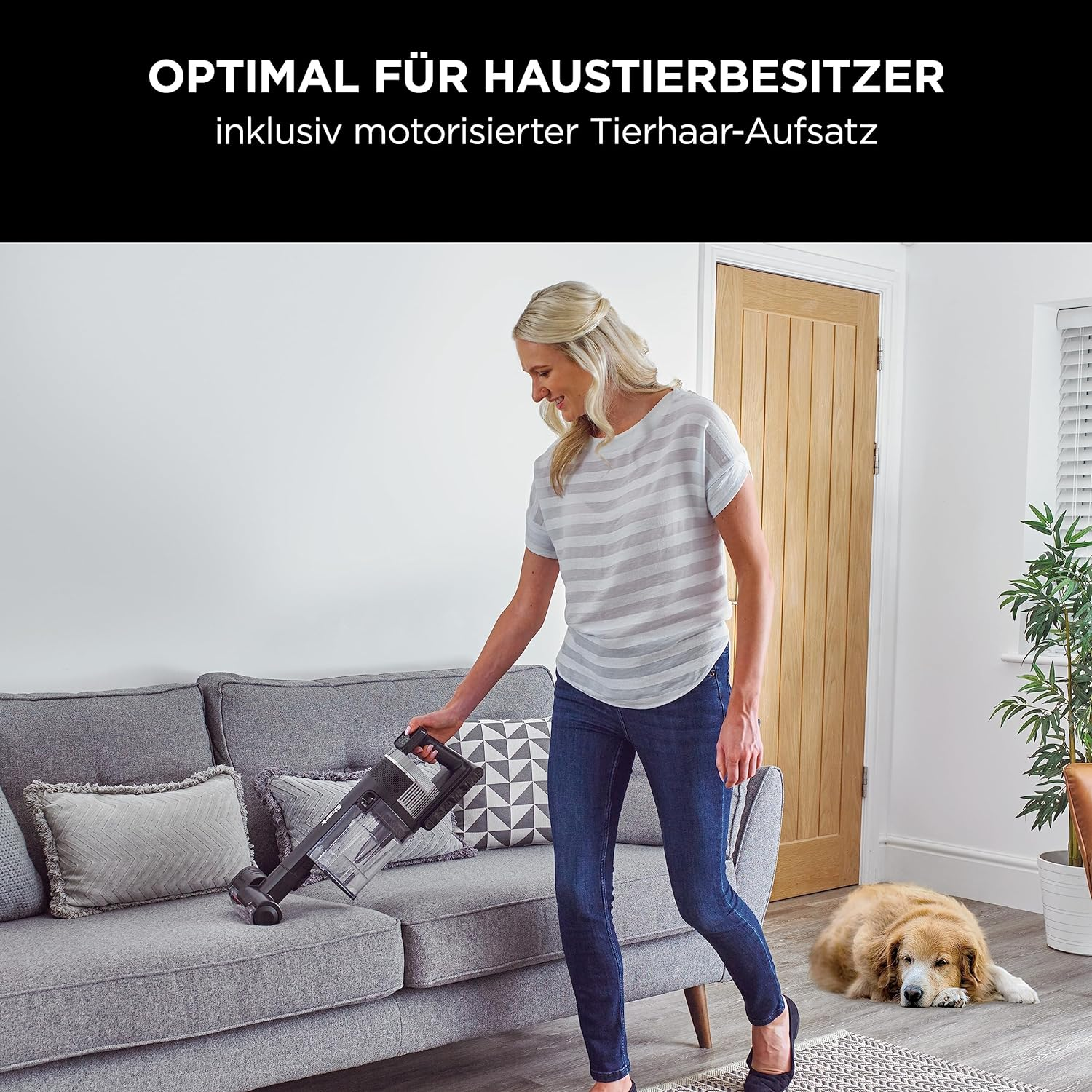 Mujer aspirando sofá gris con perro en sala de estar.