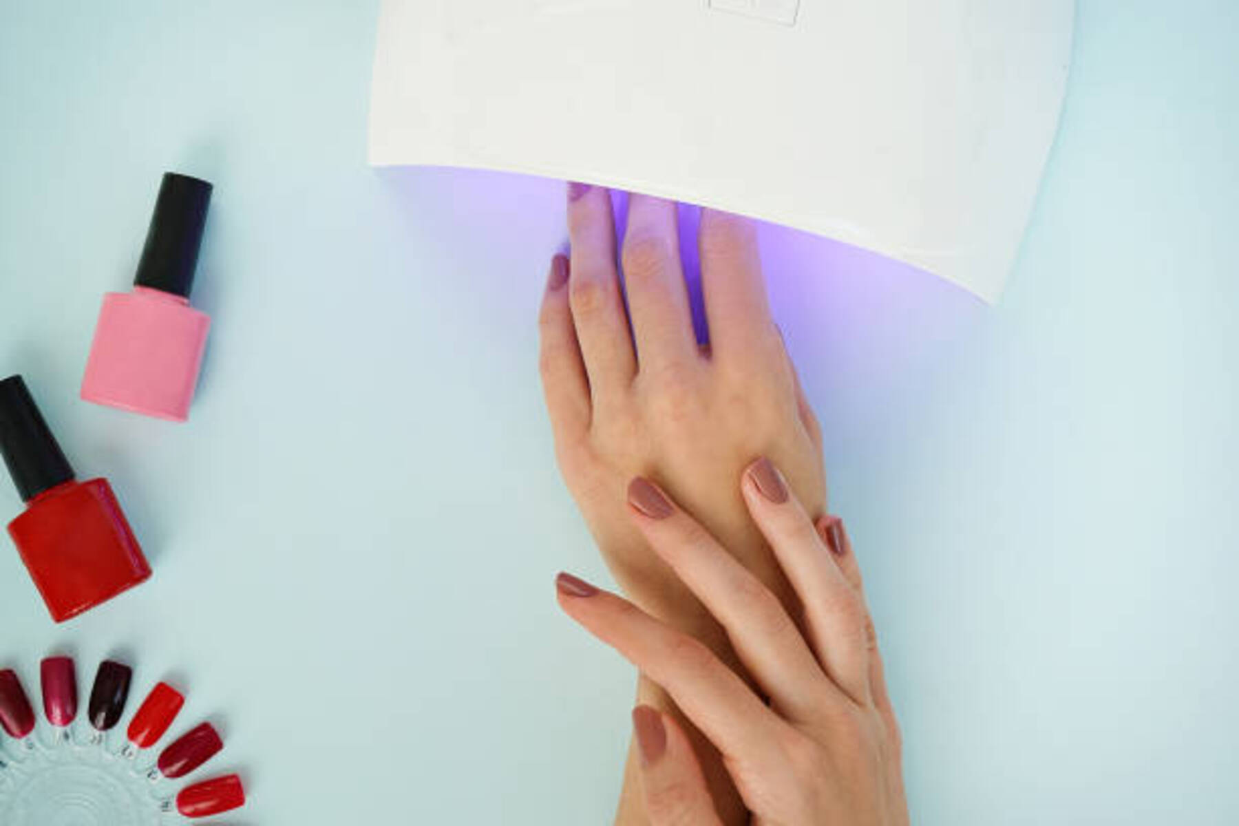 Manos bajo una lámpara UV para uñas con frascos de esmalte al lado. La escena es sobre una superficie azul claro.