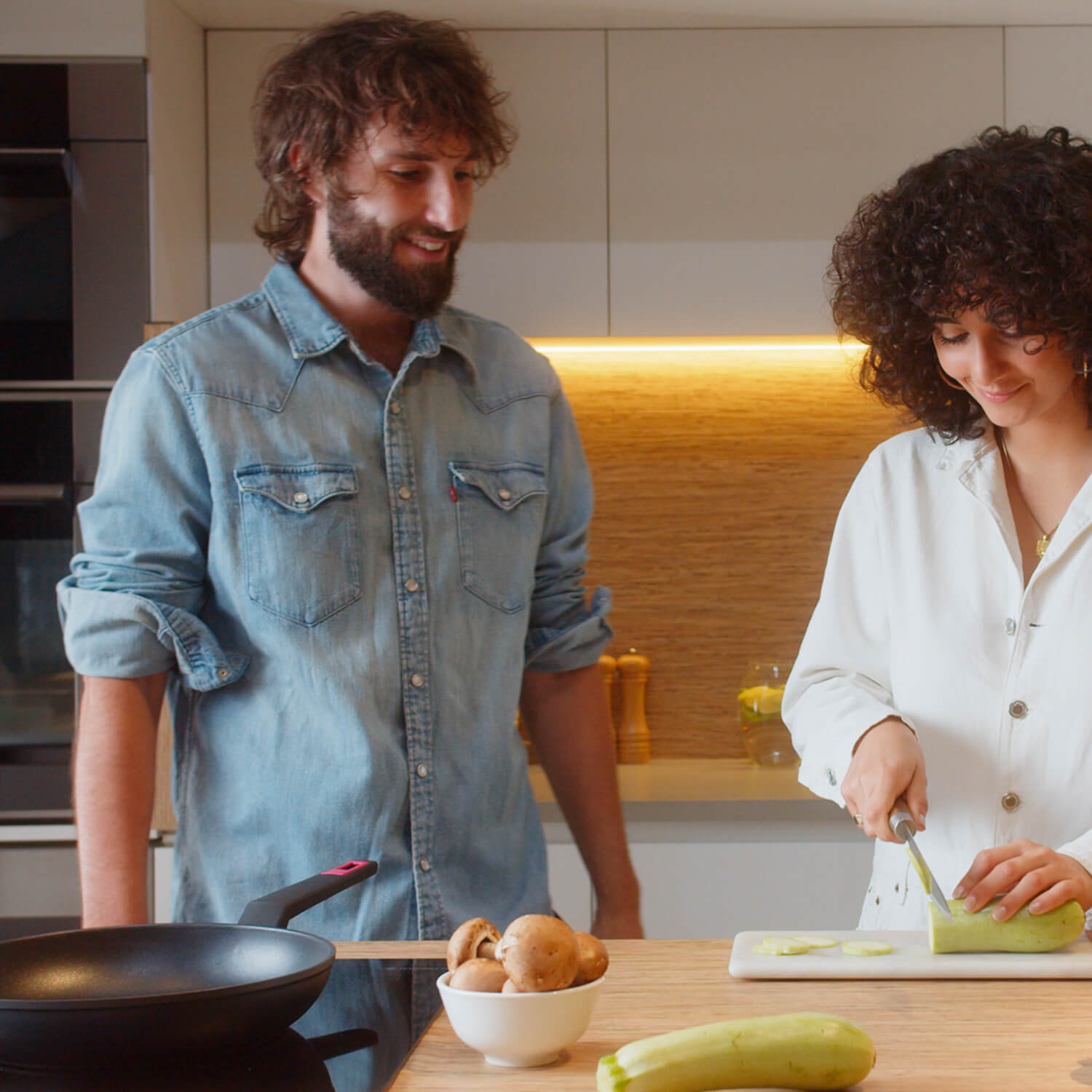 Una pareja en una cocina, uno cortando verduras, el otro mirando.
