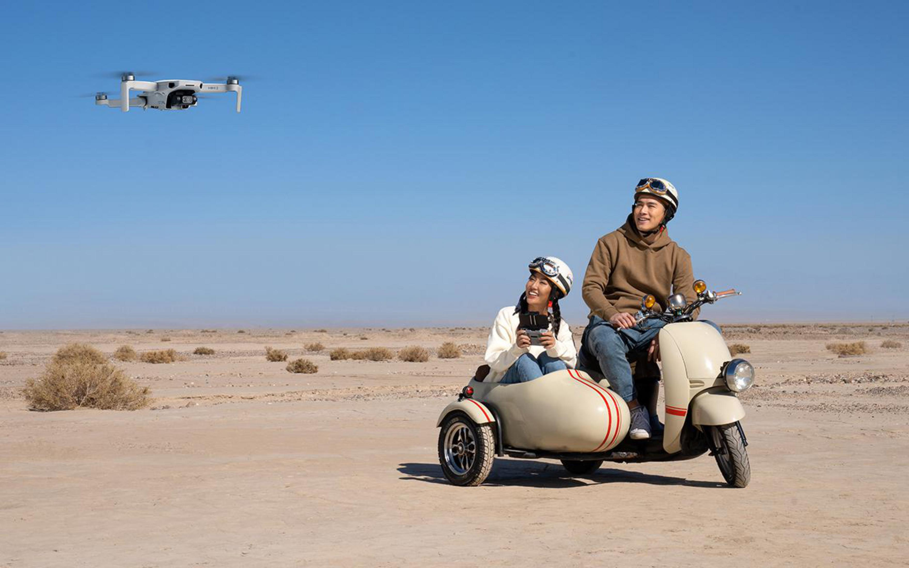 Eine Drohne fliegt über einem Paar auf einem Motorroller mit Seitenwagen in einer Wüstenlandschaft.