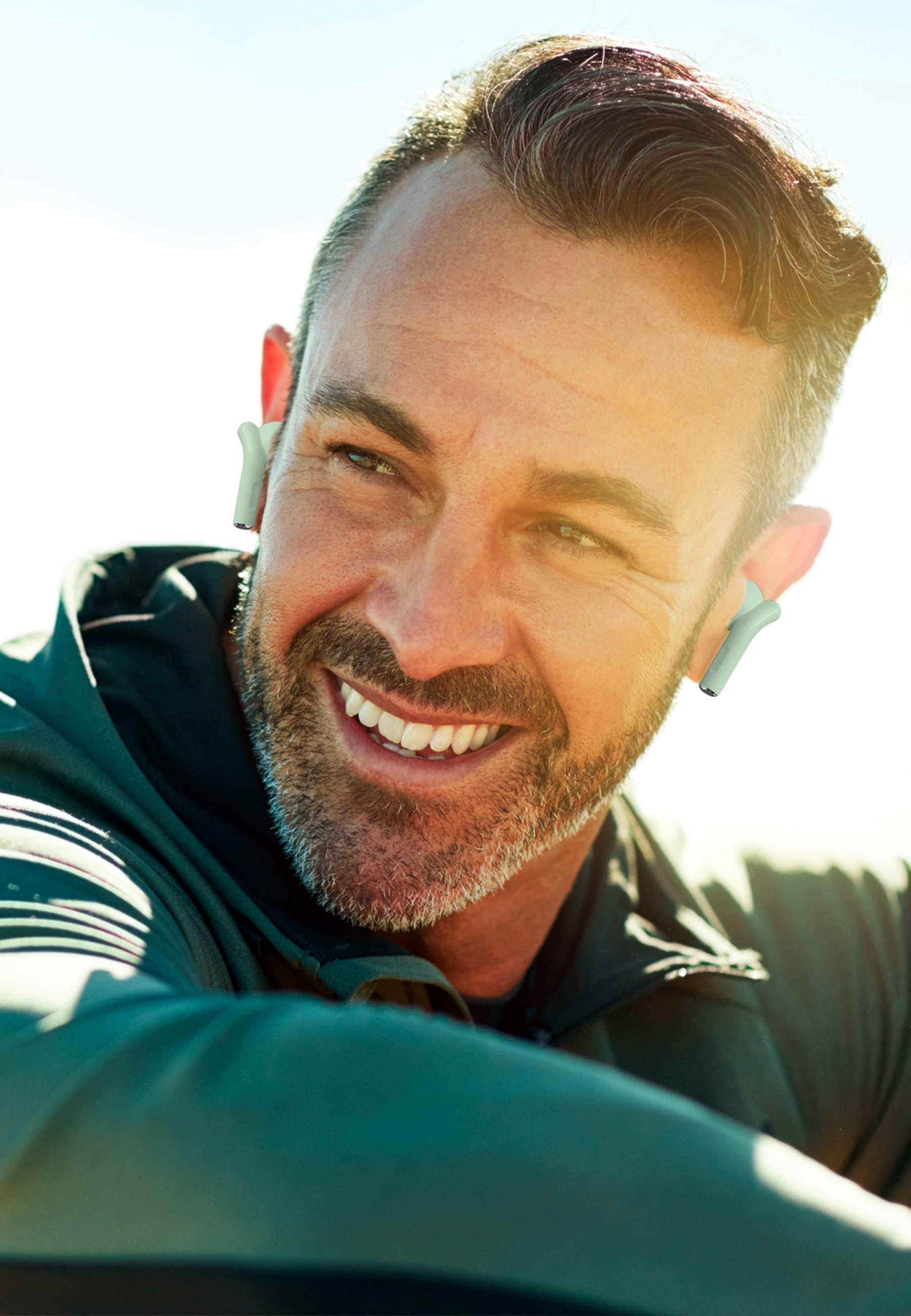 Un uomo sorridente con auricolari. Ha la barba e si trova all'aperto al sole.