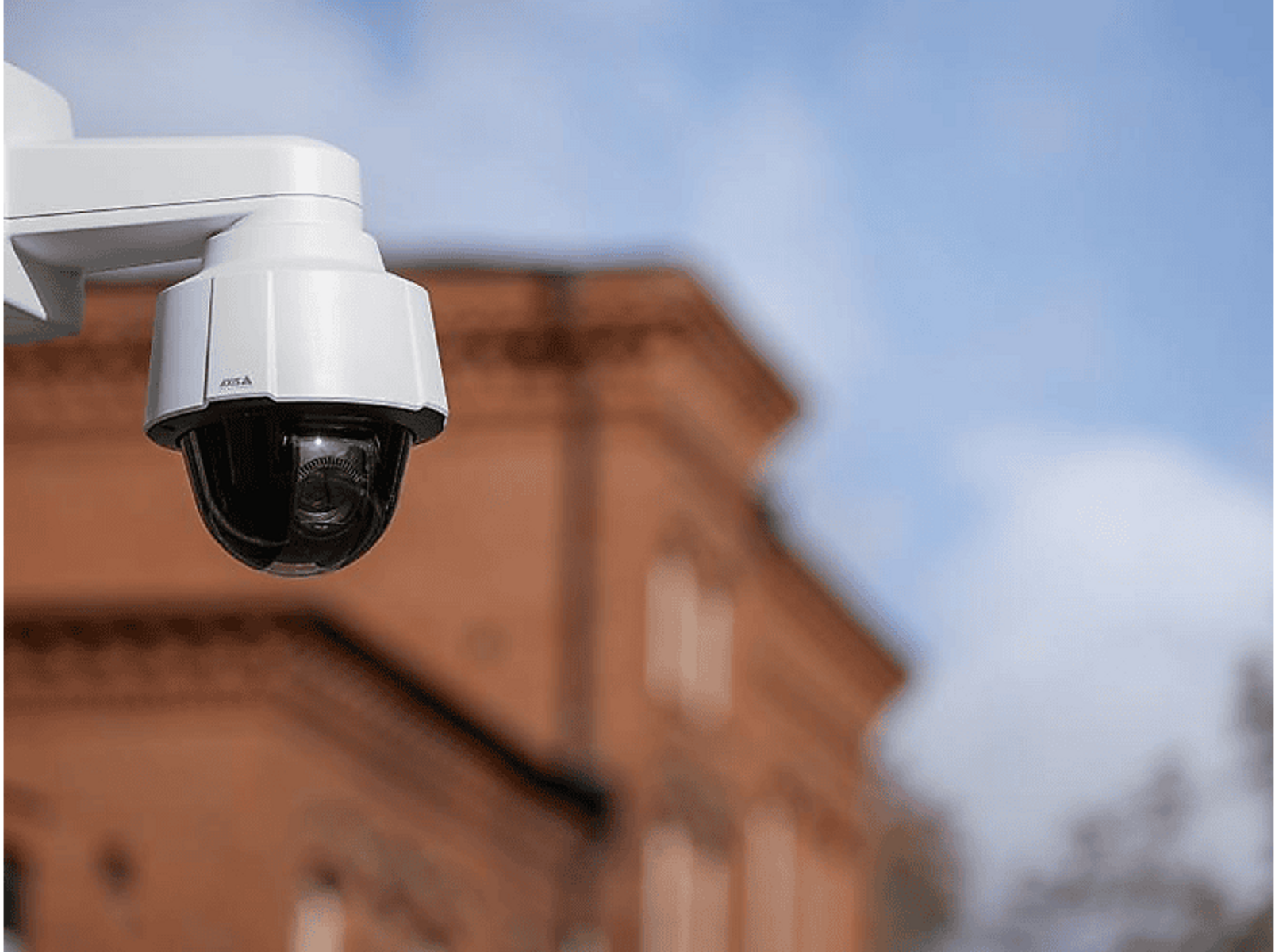 Biała kamera bezpieczeństwa na słupie. Znajduje się przed budynkiem i błękitnym niebem.