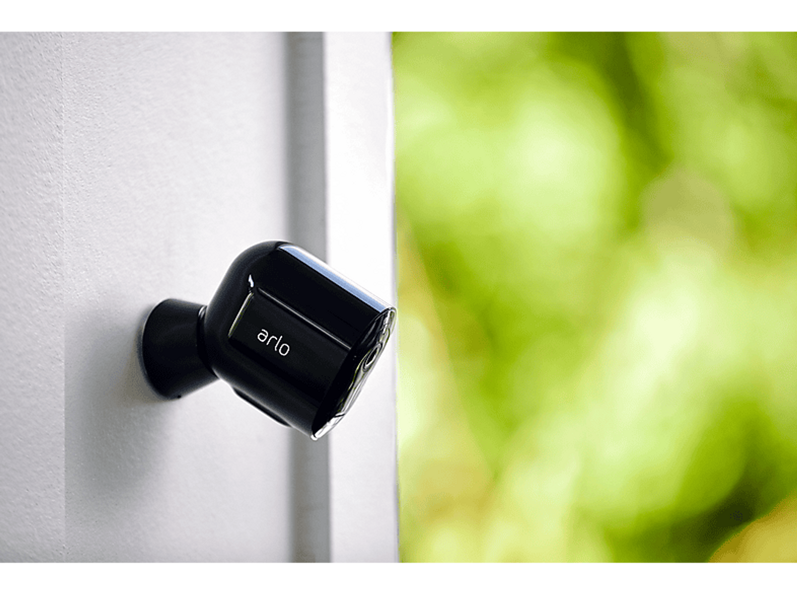 Czarna kamera bezpieczeństwa Arlo na białej ścianie, z rozmytym zielonym tłem.