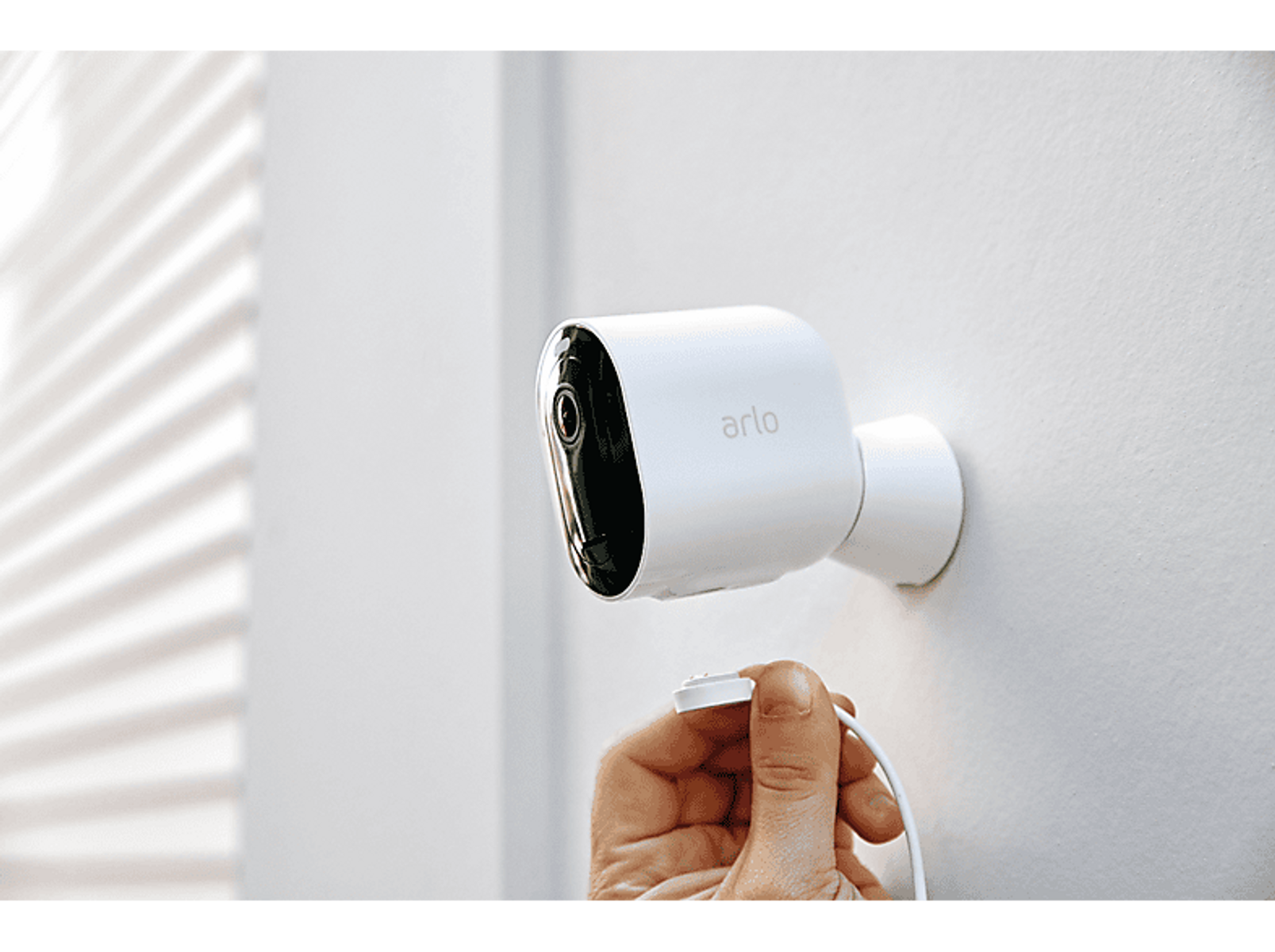 Biała kamera bezpieczeństwa Arlo zamontowana na ścianie, z kablem ładującym.