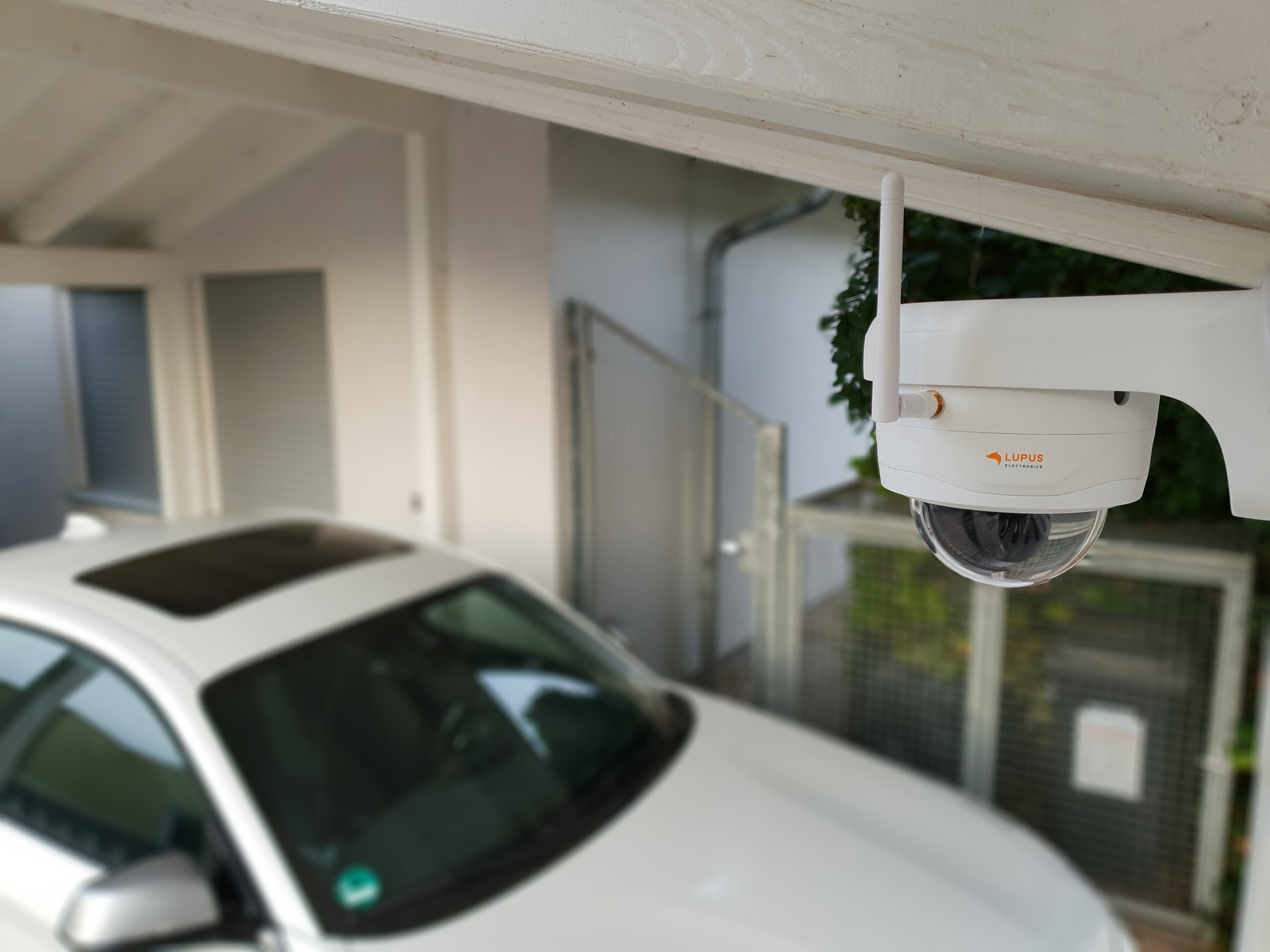 Biała kamera bezpieczeństwa jest zamontowana na zewnątrz w pobliżu białego samochodu.