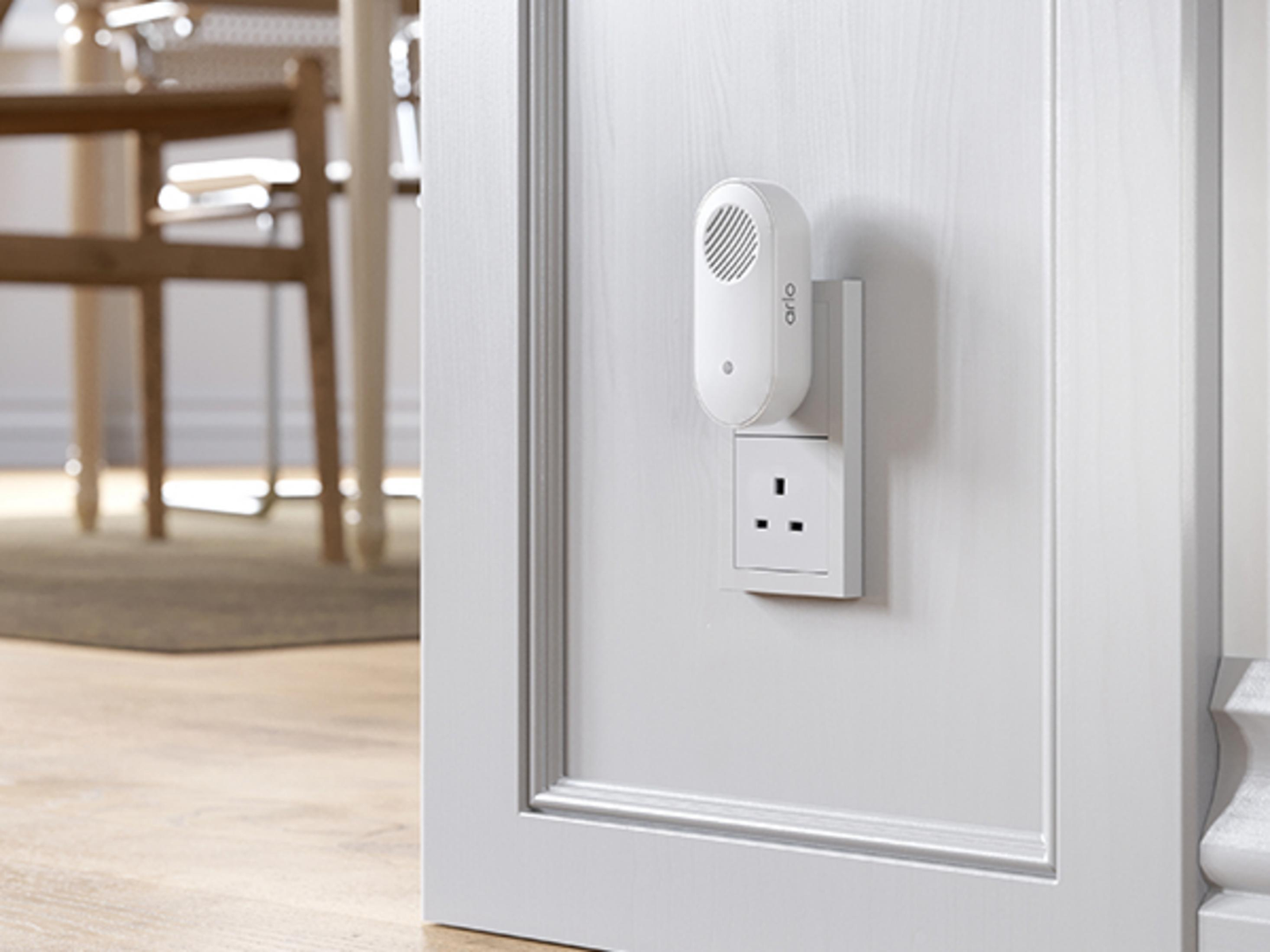 Un dispositivo di sicurezza Arlo bianco collegato a una presa a muro su un telaio di porta bianco.