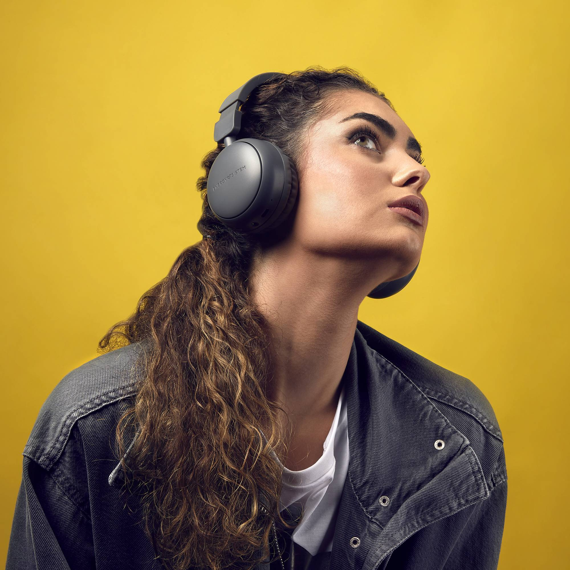 Mujer con auriculares grises, mirando hacia arriba. Fondo amarillo. Cabello oscuro y chaqueta de mezclilla.