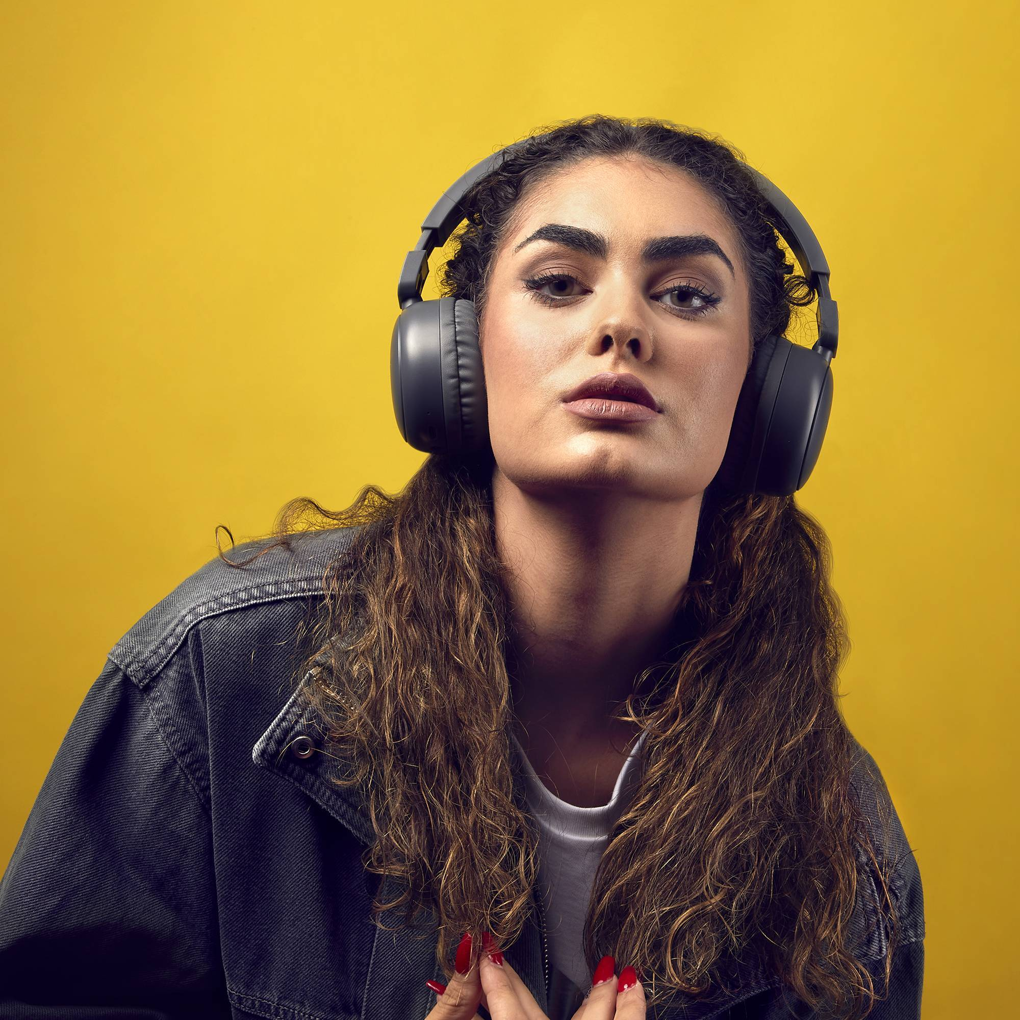 Mujer con auriculares. Tiene cabello castaño y esmalte de uñas rojo. Fondo amarillo.