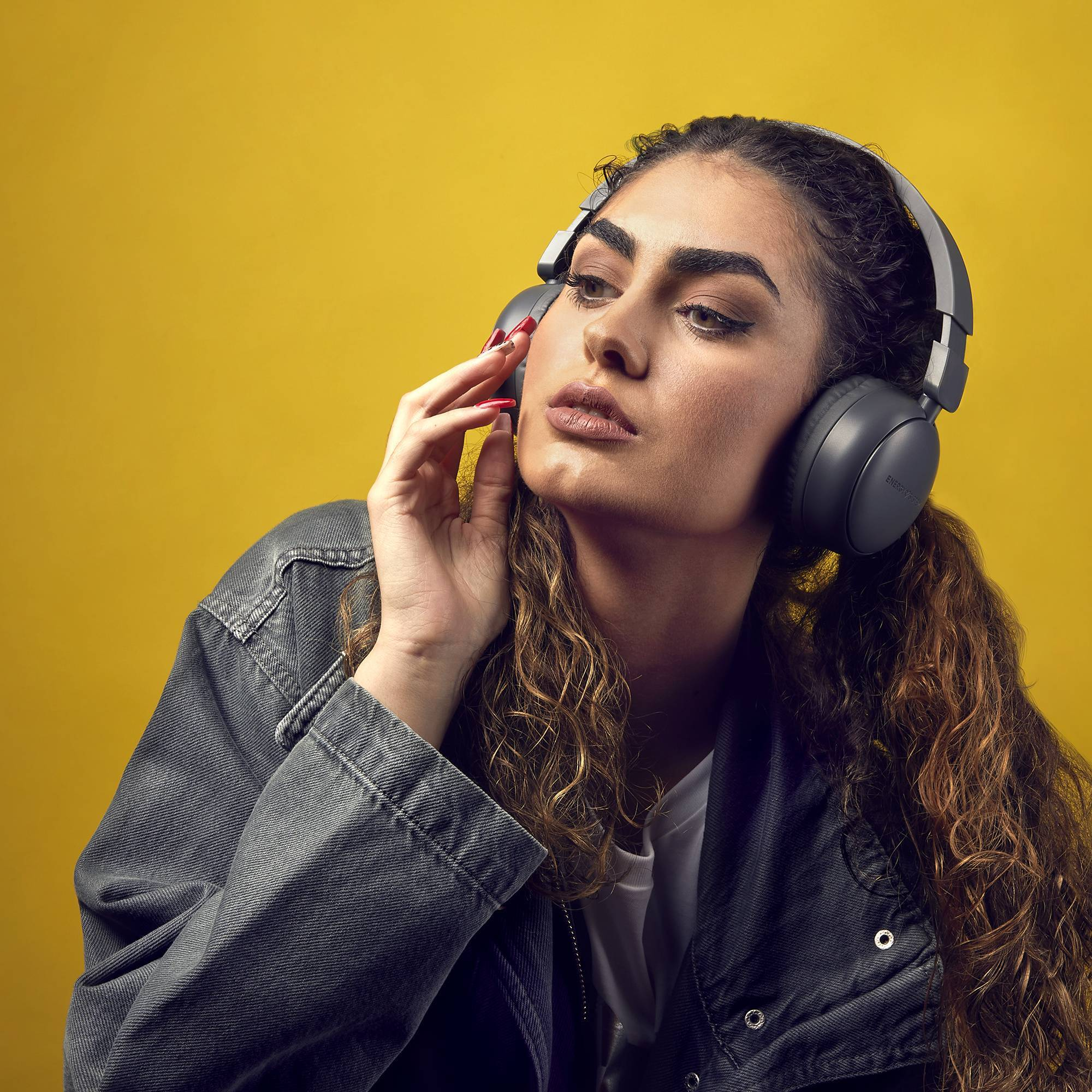 Mujer con auriculares sobre un fondo amarillo, sosteniendo su mano en su rostro.