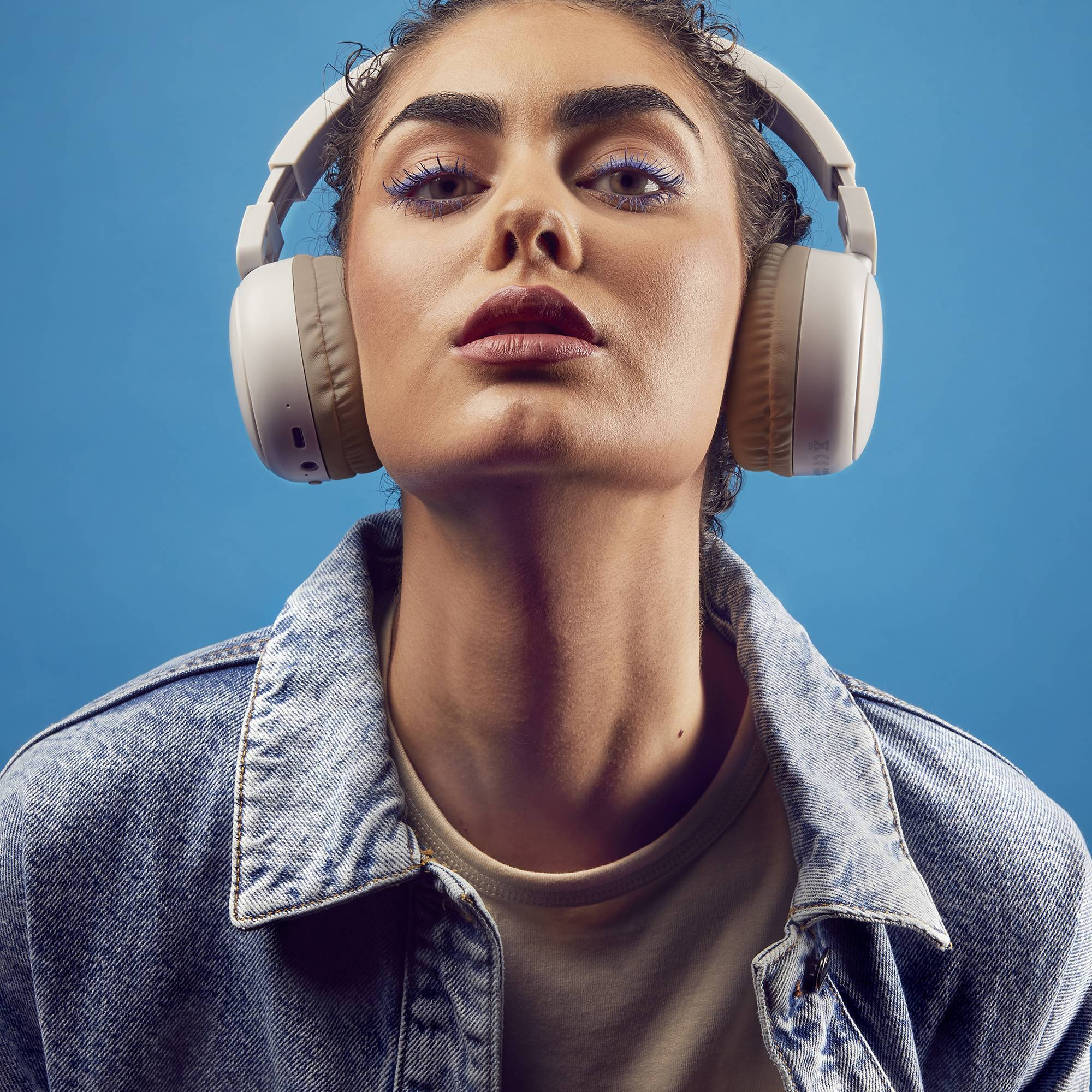 Kobieta w białych słuchawkach, dżinsowej kurtce, patrząca w górę na niebieskim tle.