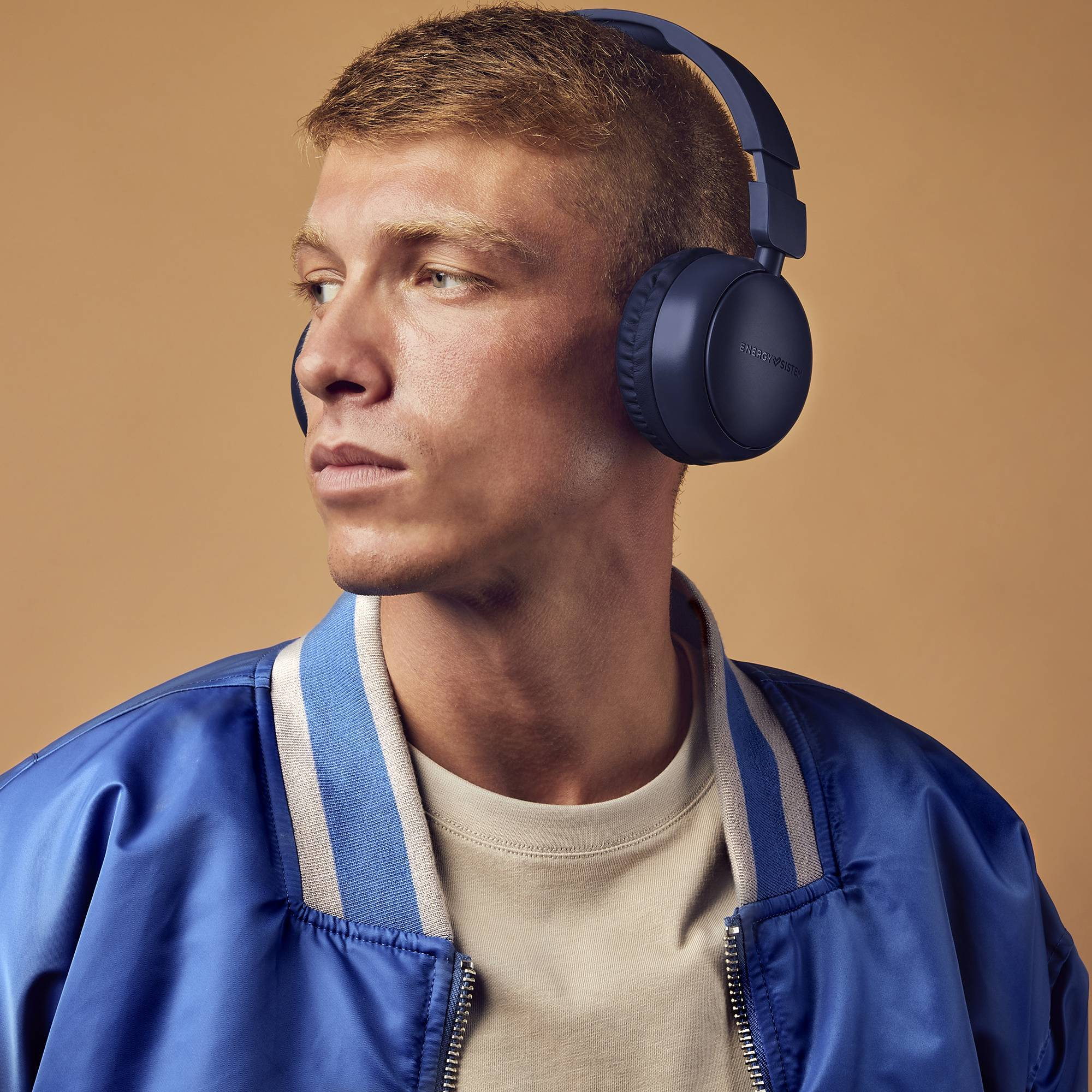 Ein Mann mit blauen Kopfhörern und einer blauen Jacke, der nach links schaut, hellbrauner Hintergrund.