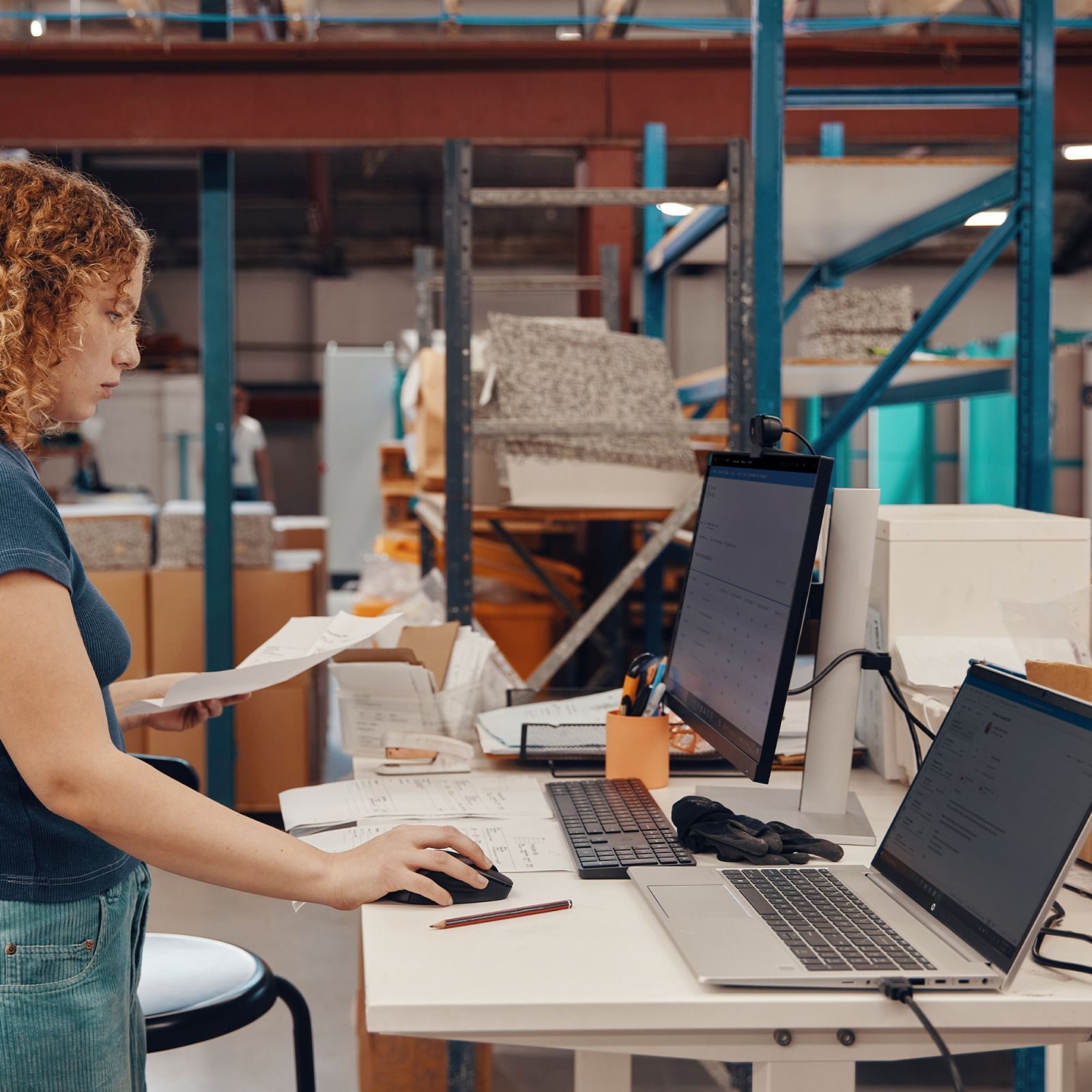 Kobieta pracująca na laptopie z myszką i papierami w magazynie.