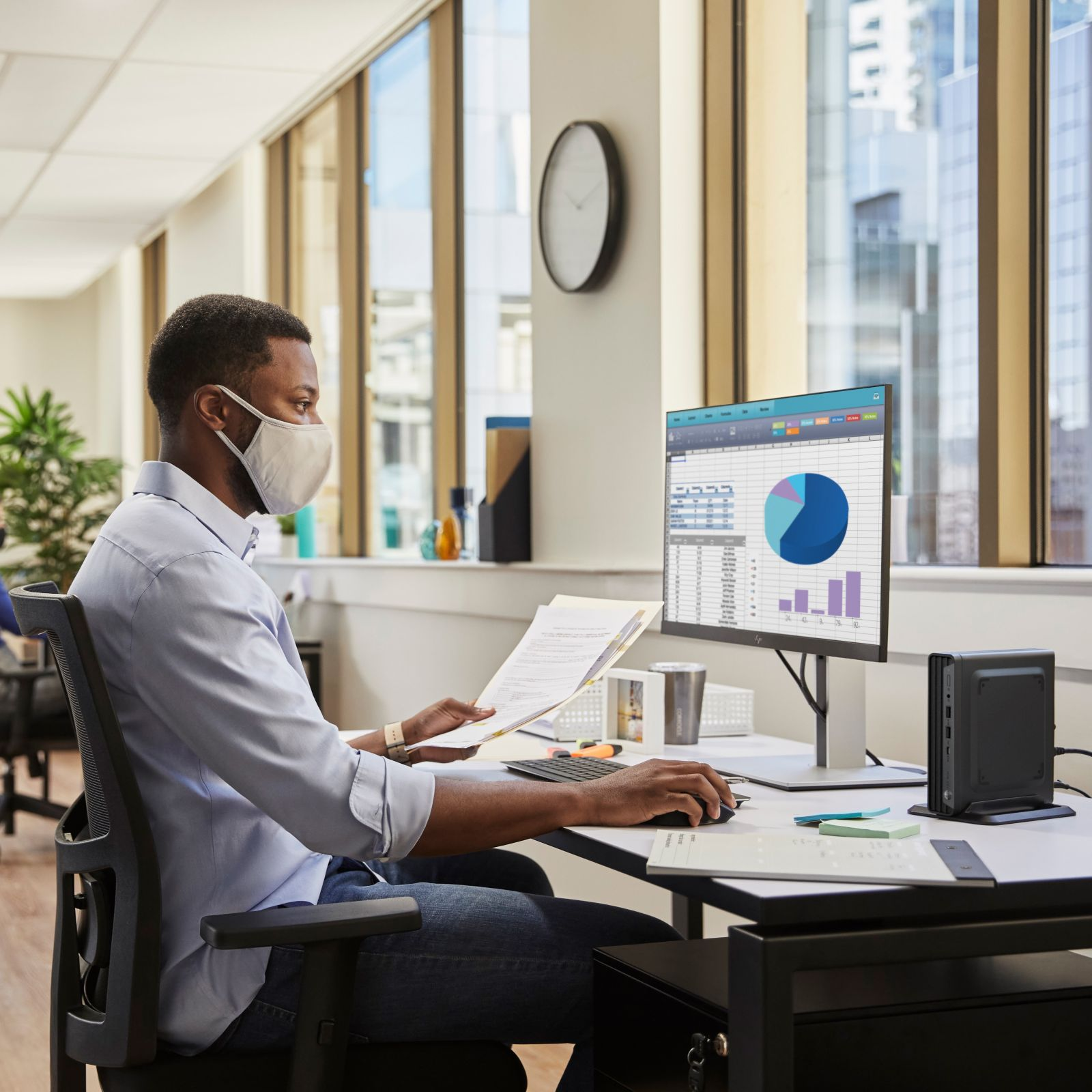 Mężczyzna w masce przy biurku patrzący na monitor. Papiery i klawiatura na biurku. Okna w tle.