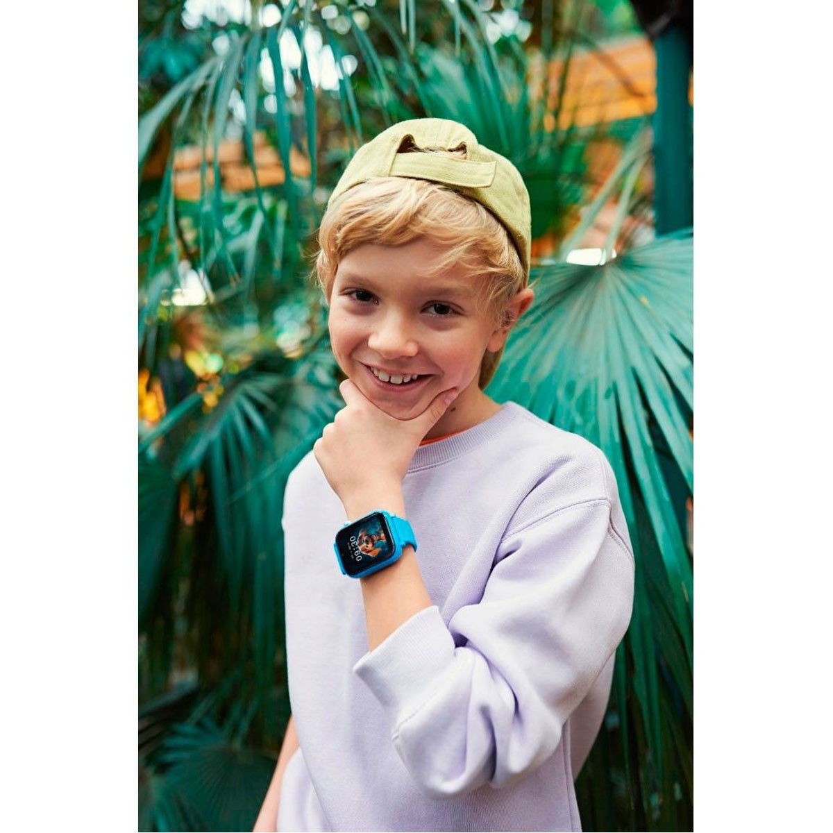 Jongen met een blauwe smartwatch glimlacht terwijl hij zijn kin vasthoudt. Heeft een groene hoed en een lichtpaars shirt.