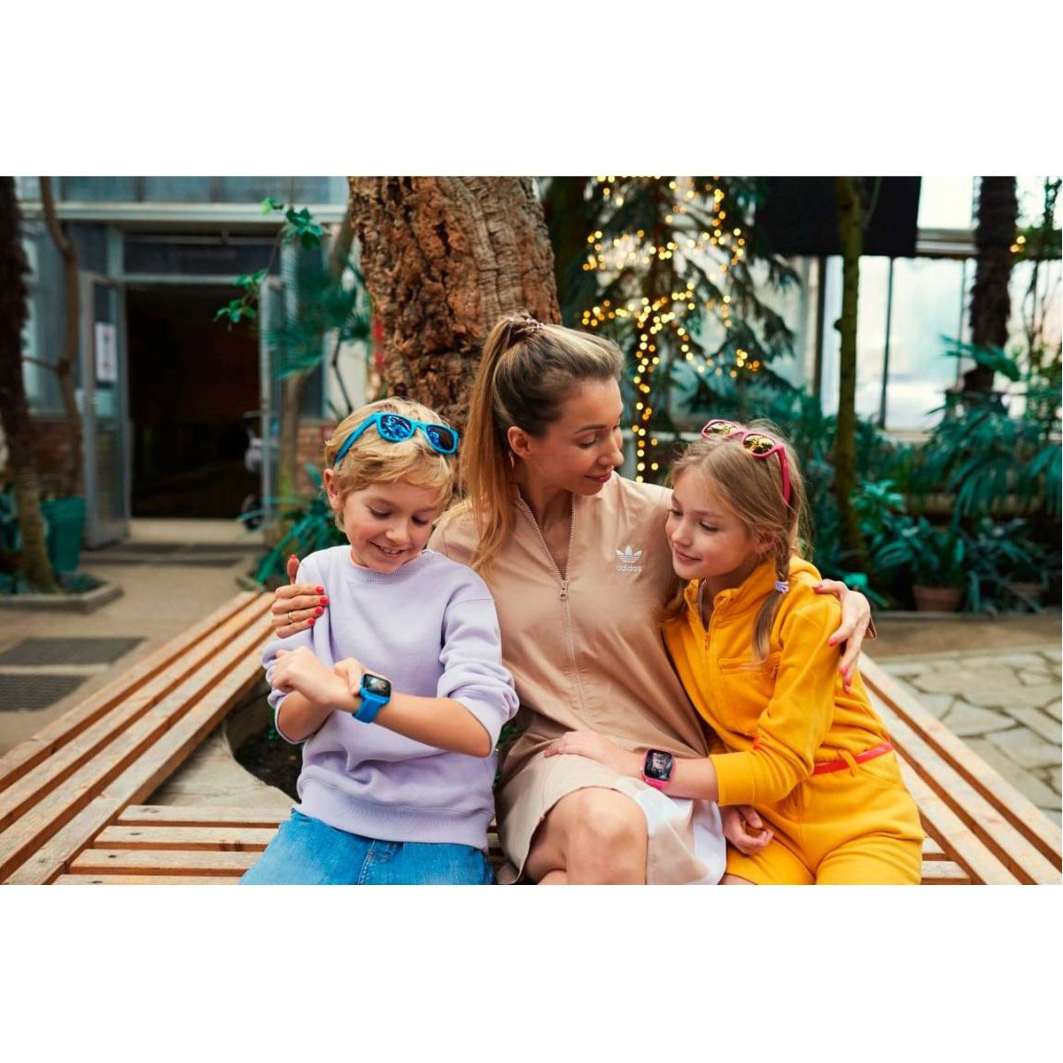 Een vrouw en twee kinderen zitten op een houten bankje, ze dragen allemaal smartwatches en lachen.