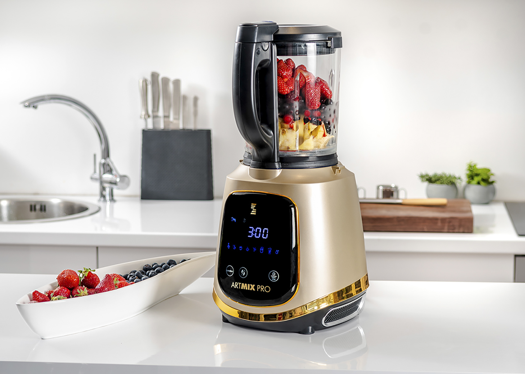 Ein goldfarbener Mixer mit Früchten im Inneren, auf einer weißen Arbeitsplatte. Eine Schüssel Beeren ist vorne.