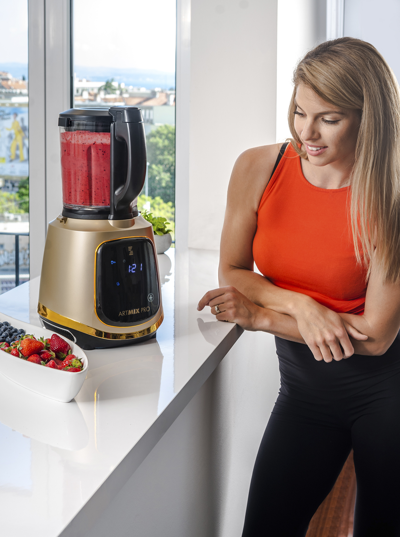 Frau bereitet Smoothie in goldenem Mixer zu, Zutaten auf Theke, heller Fensterblick.