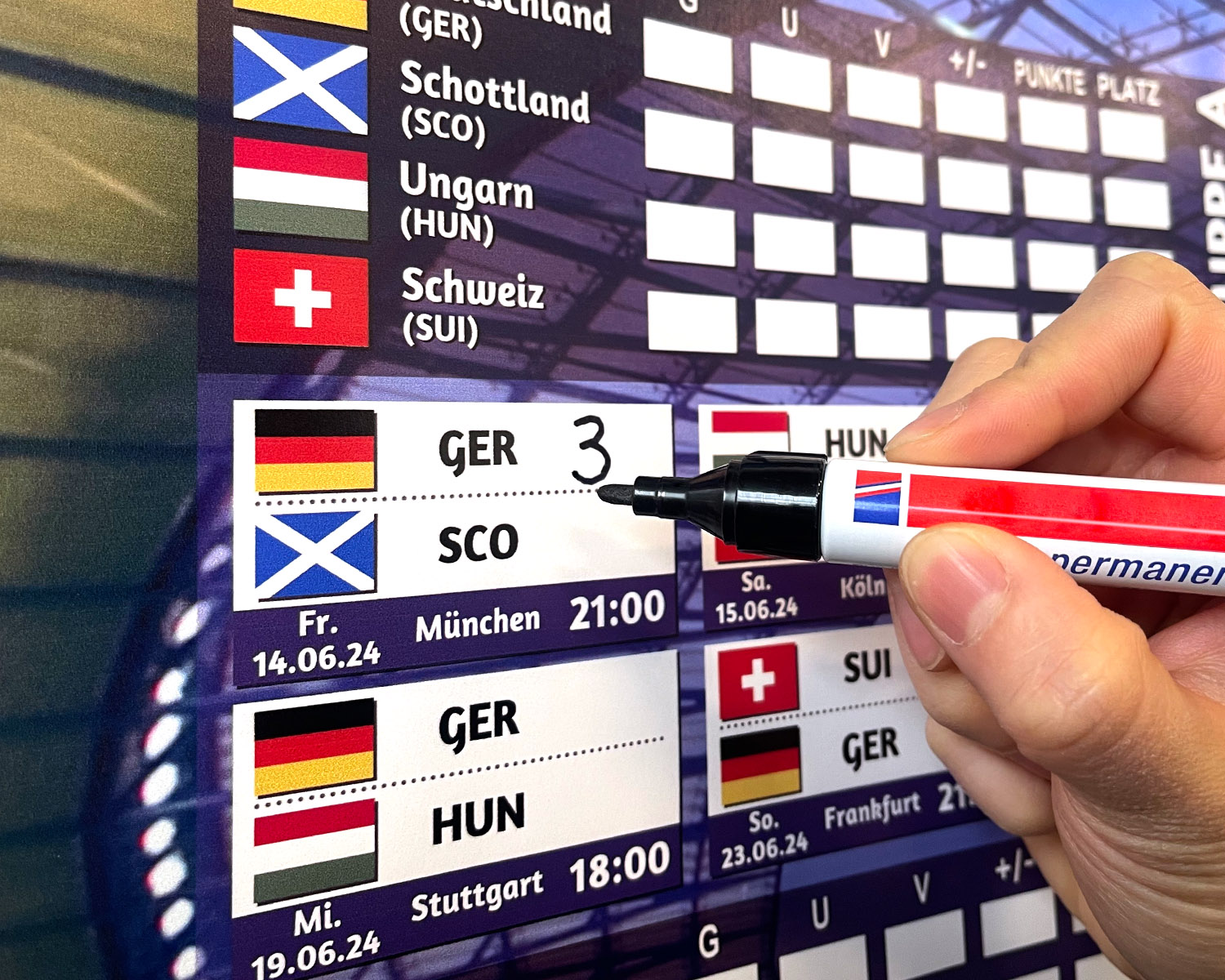Hand schreibt GER 3 auf eine Spielplan-Tafel mit Flaggen von Deutschland und Schottland.