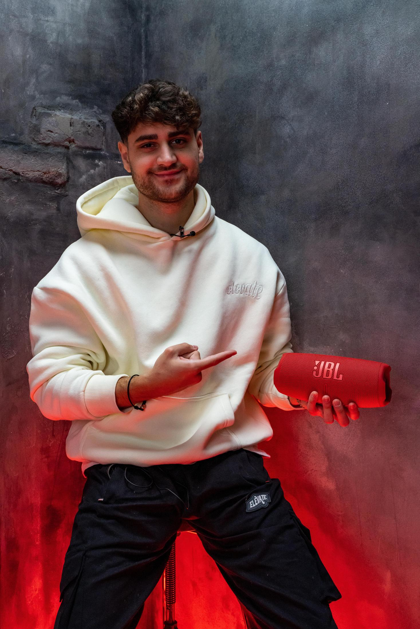 Mężczyzna w bluzie z kapturem wskazuje na czerwony głośnik JBL, uśmiechając się, na ciemnym tle.