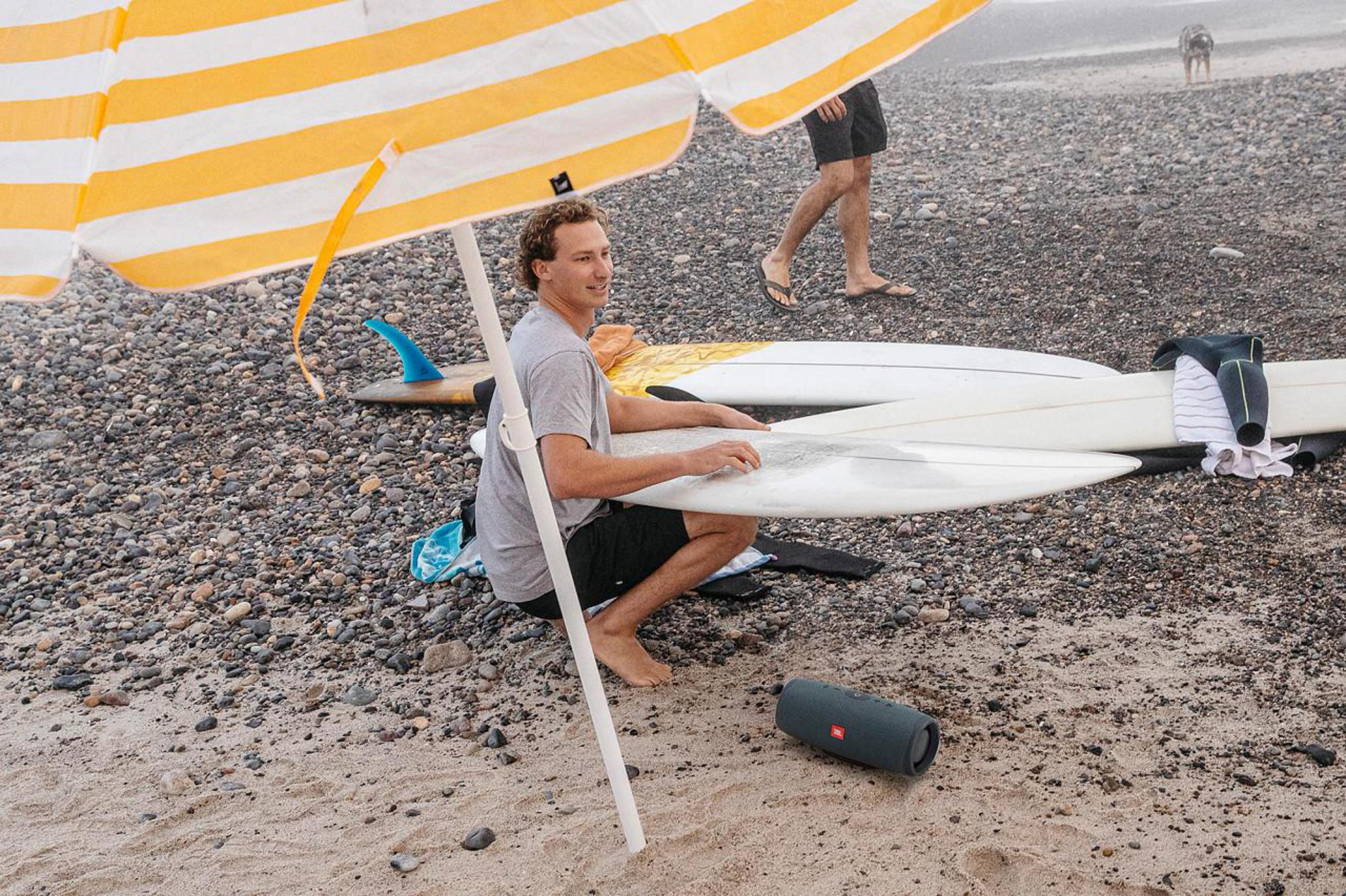 Mężczyzna z deską surfingową pod parasolem w paski, na plaży.