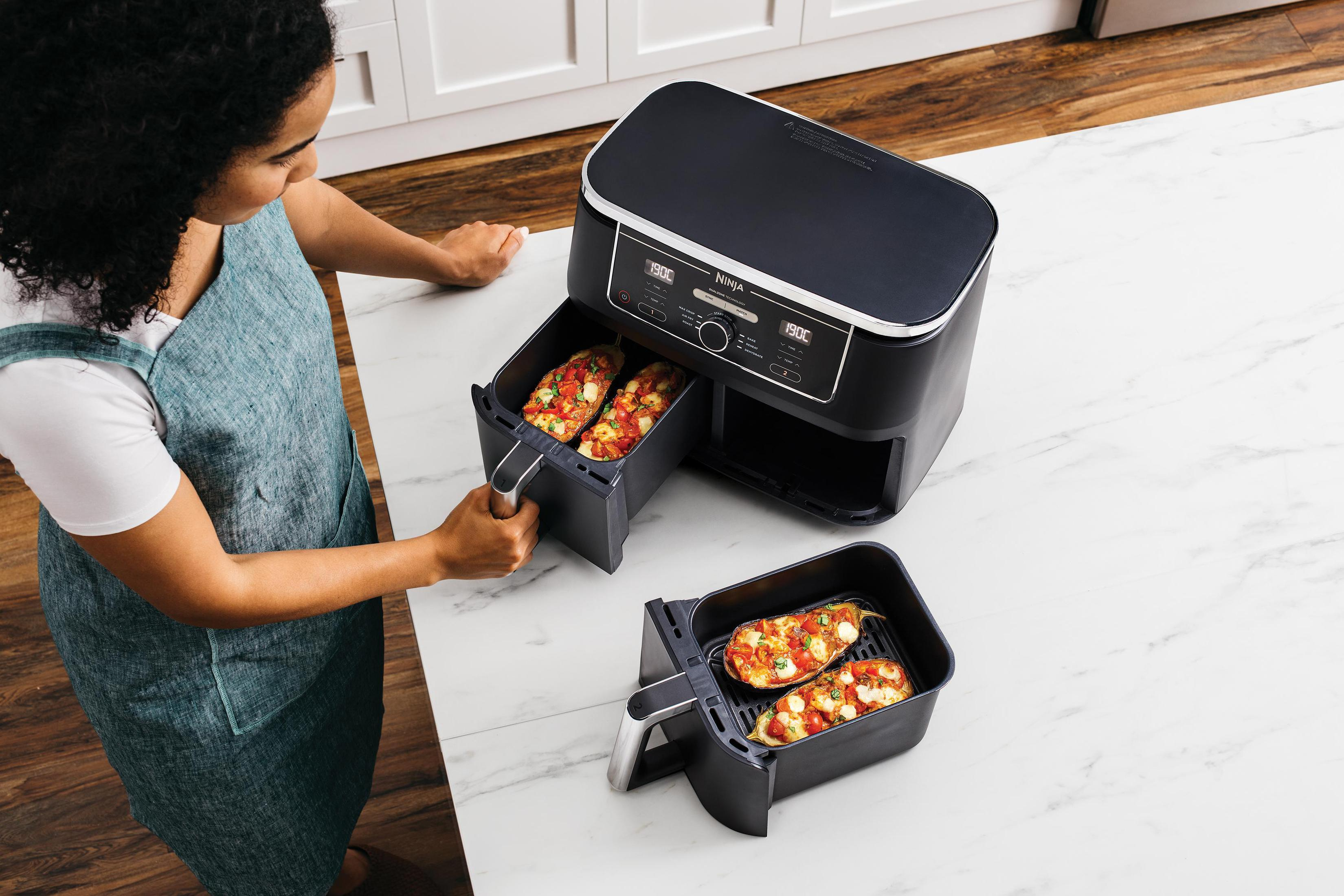 Kobieta używa frytkownicy beztłuszczowej. Frytkownica jest czarna, są dwie tacki z jedzeniem.