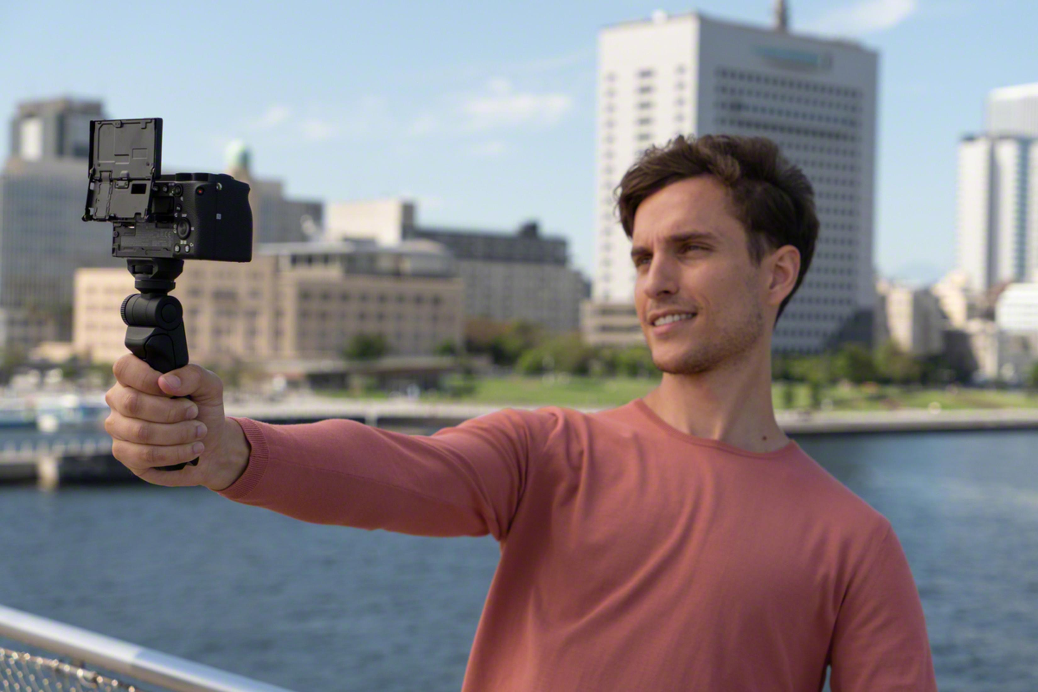 Un hombre sonriendo, sosteniendo una cámara con una pantalla abatible, con edificios de la ciudad al fondo.
