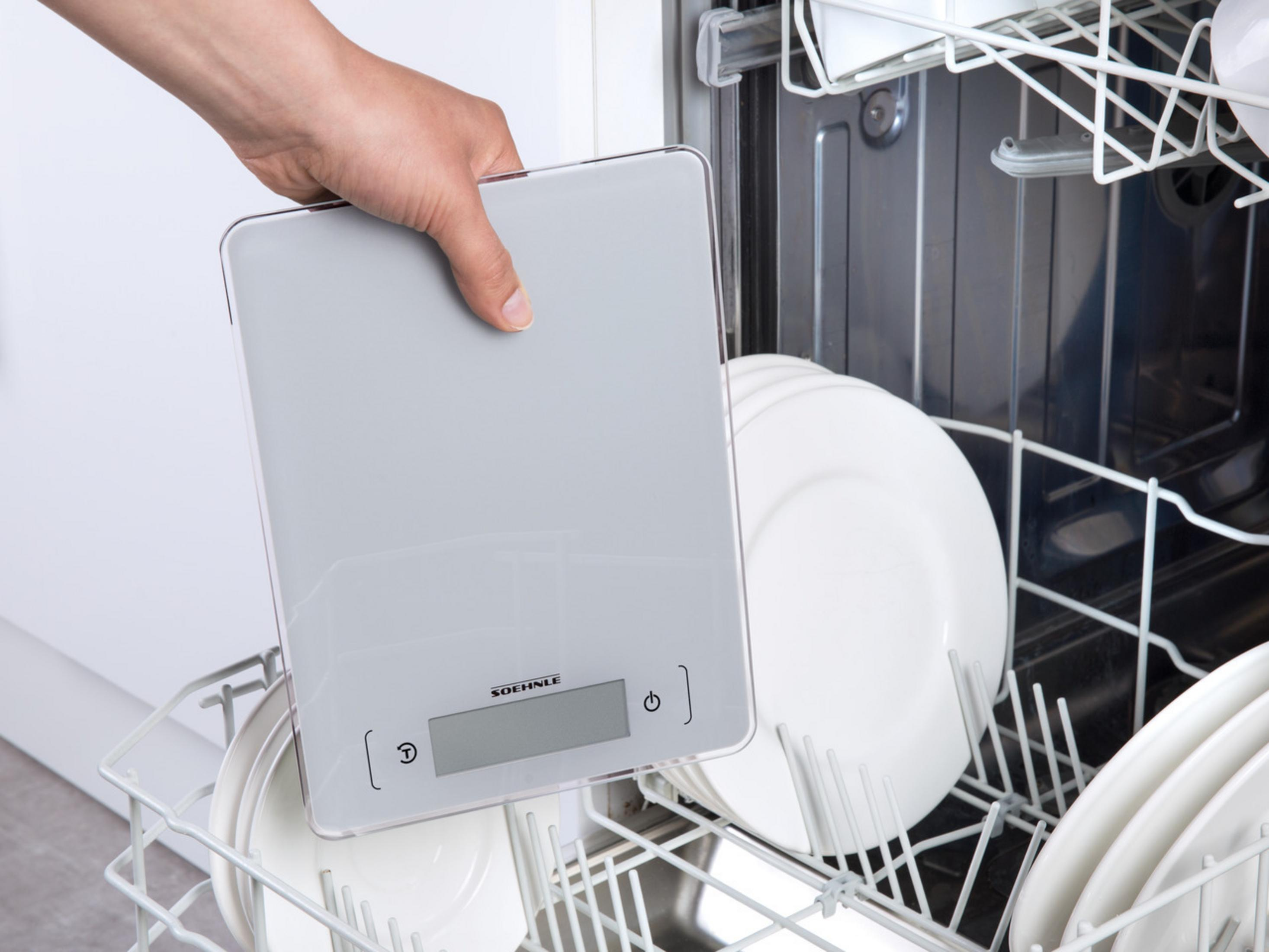 Une main tenant une balance de cuisine numérique dans un lave-vaisselle. Des assiettes blanches sont sur le support.