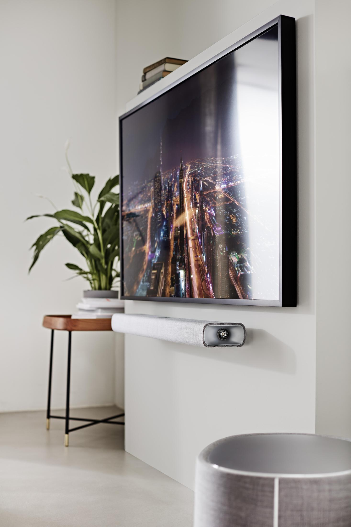 Ein Fernseher an der Wand mit einer Soundbar. Ein kleiner Tisch und eine Pflanze links.