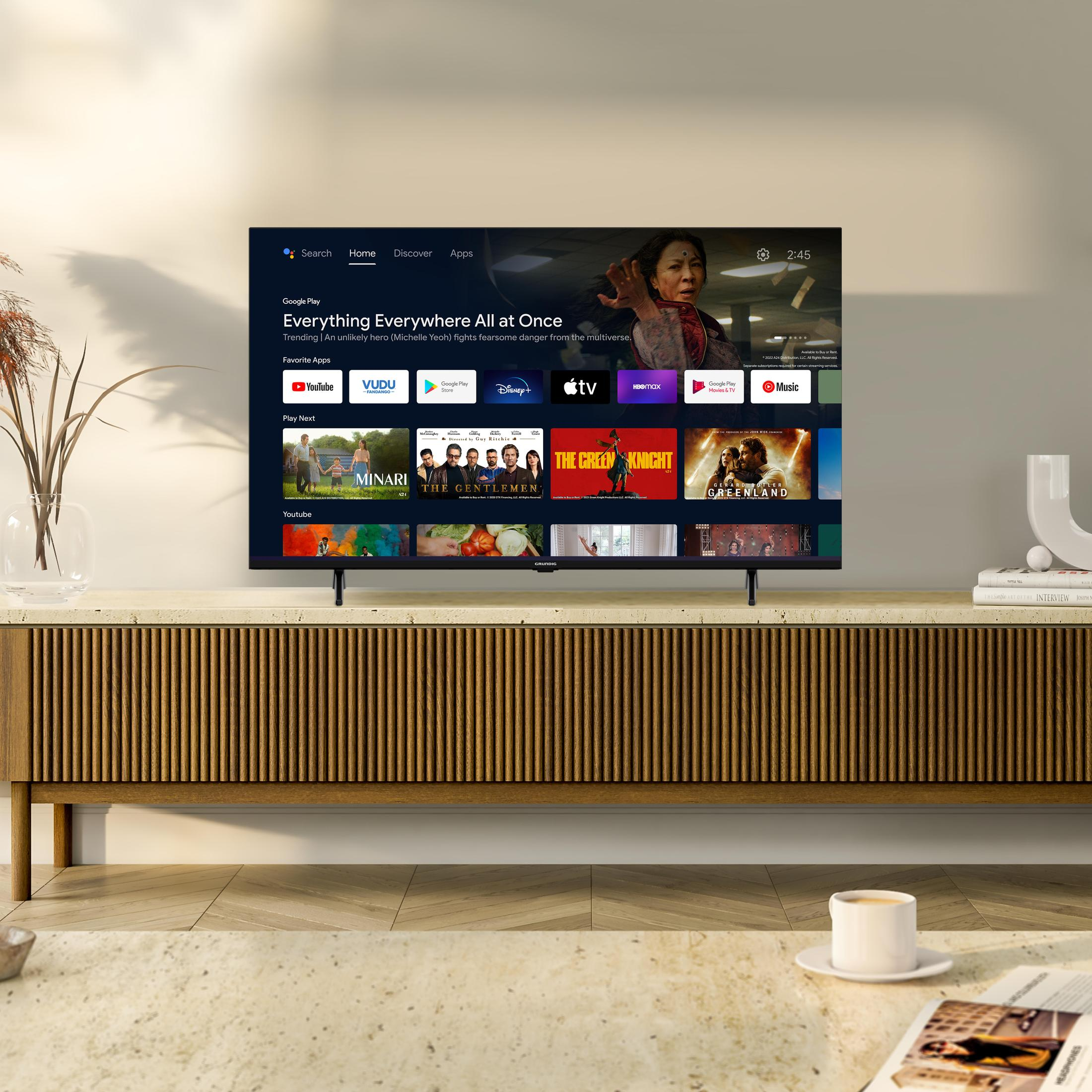 Ein Fernseher zeigt eine Filmauswahl. Der Fernseher steht auf einem Holzschrank mit einer Vase und einer Pflanze.