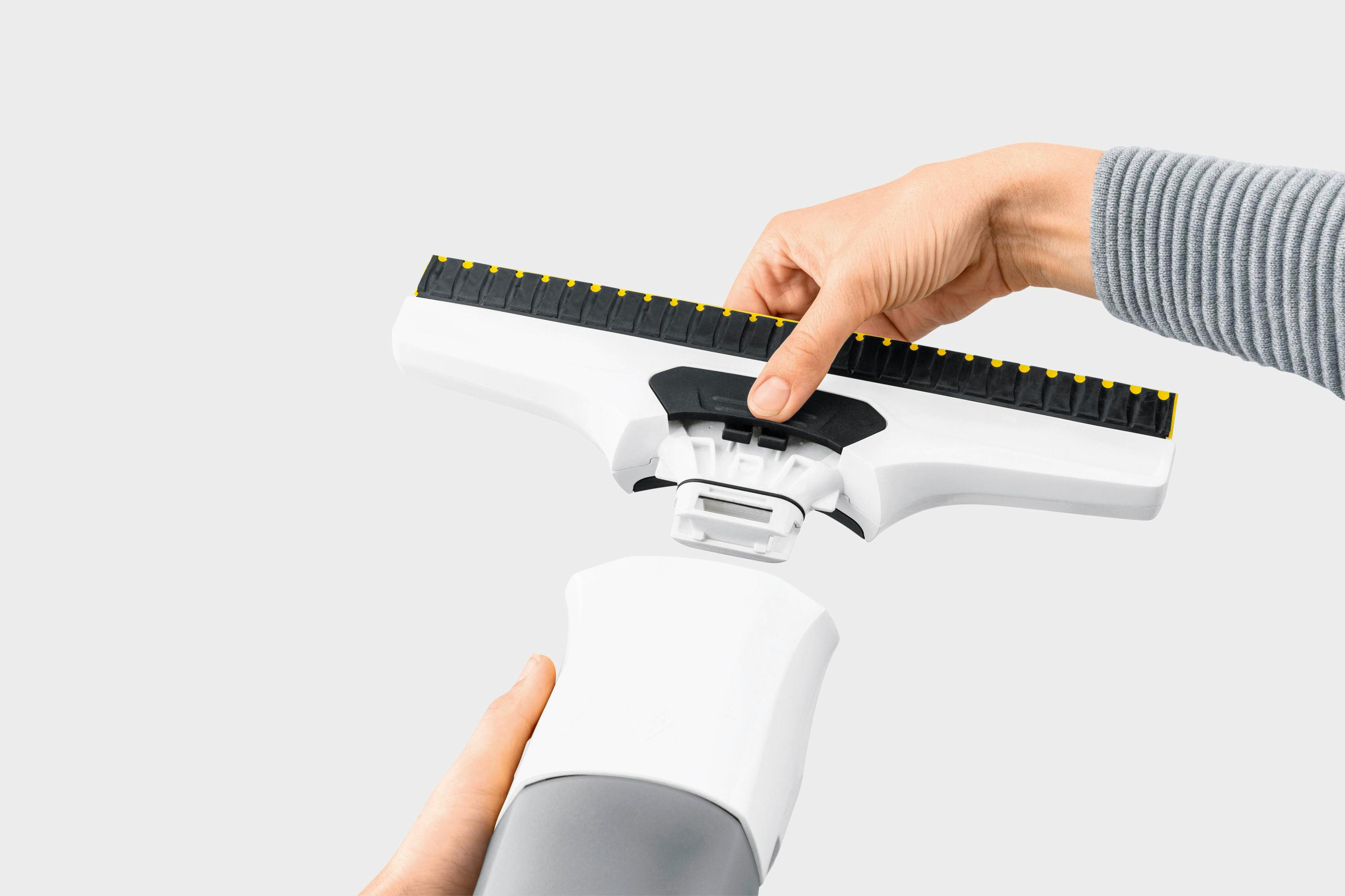 Ręce montujące biało-czarną myjkę do okien. Tło jest gładko białe.