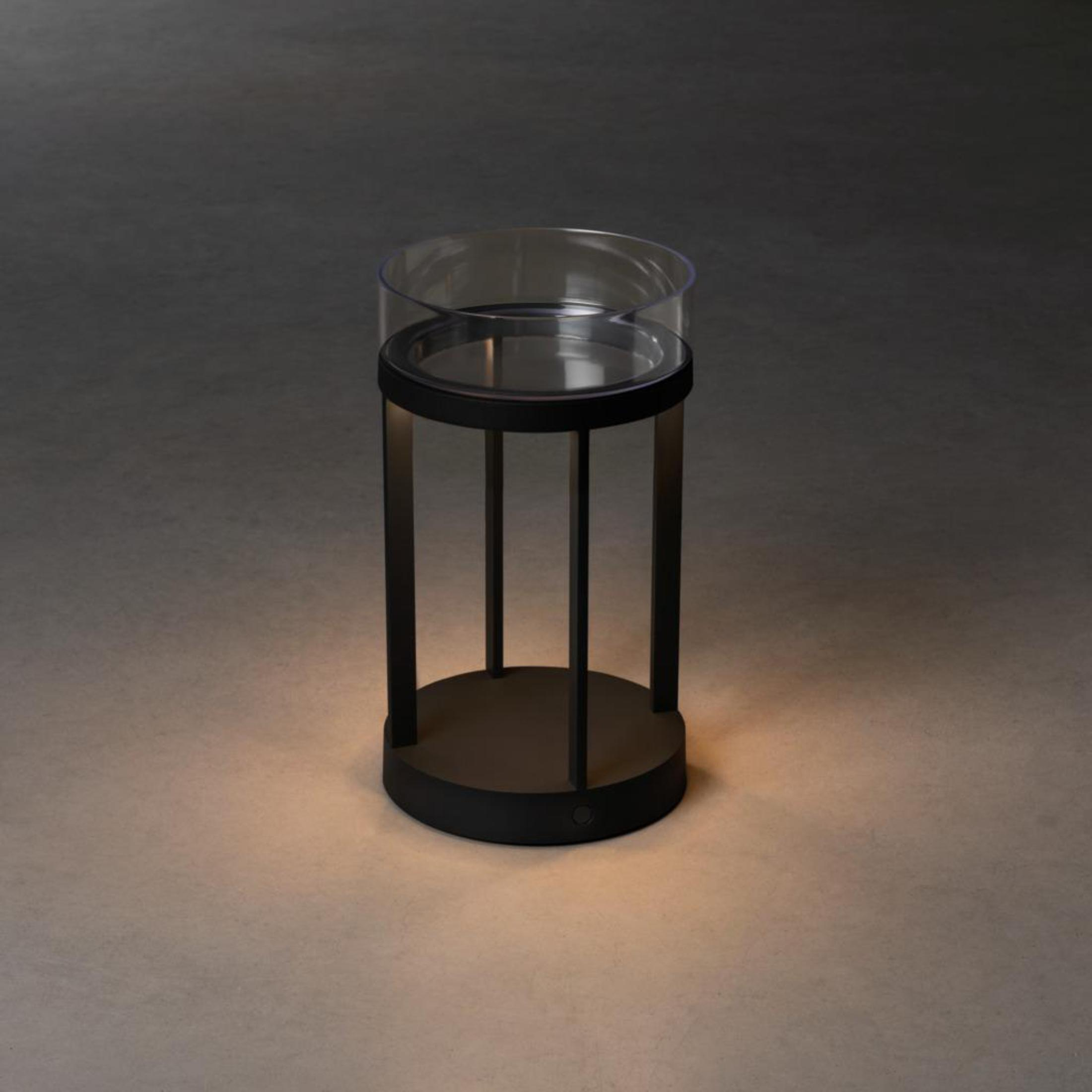 Schwarze zylindrische Lampe mit Glasaufsatz auf dunklem Boden.