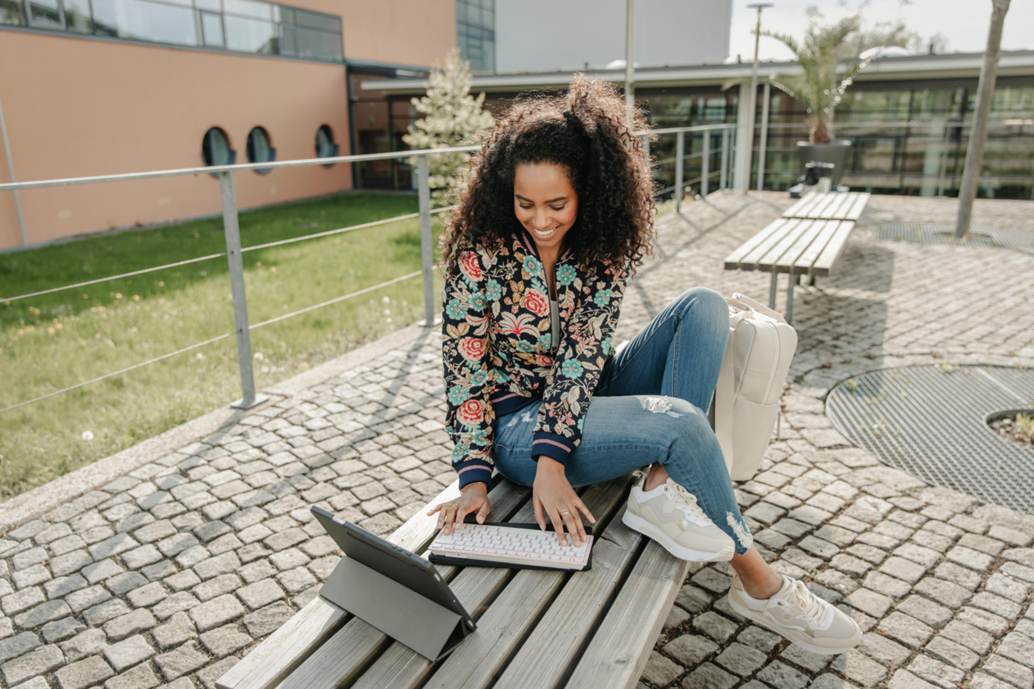 Woman sitting on a bench, working on a tablet with a keyboard. She's smiling, with backpack, outdoors in sunlight.