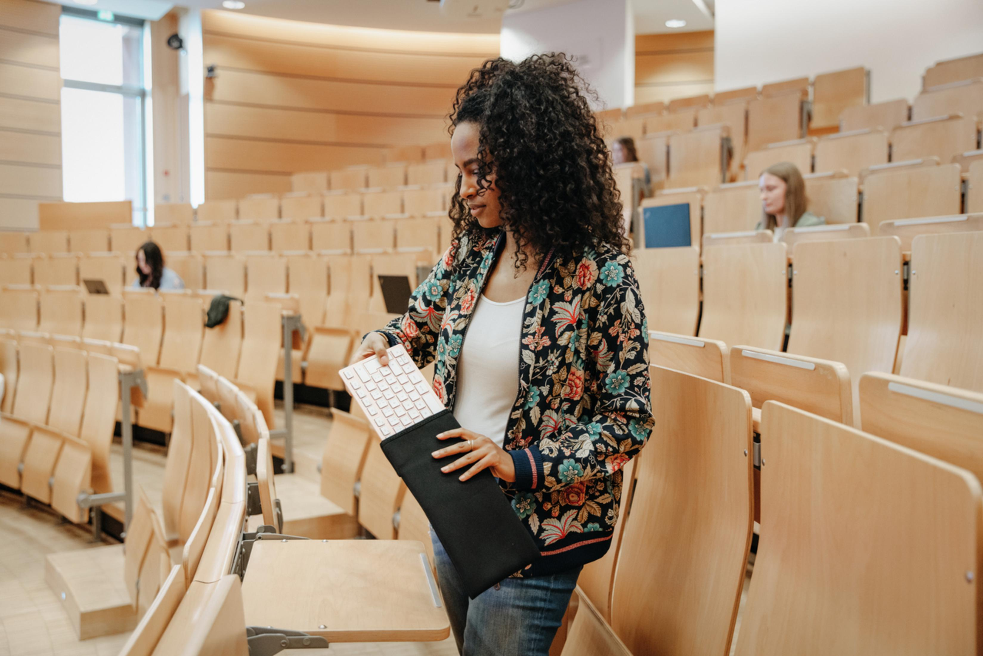 A woman in a floral jacket is putting a keyboard into a black case in an auditorium.