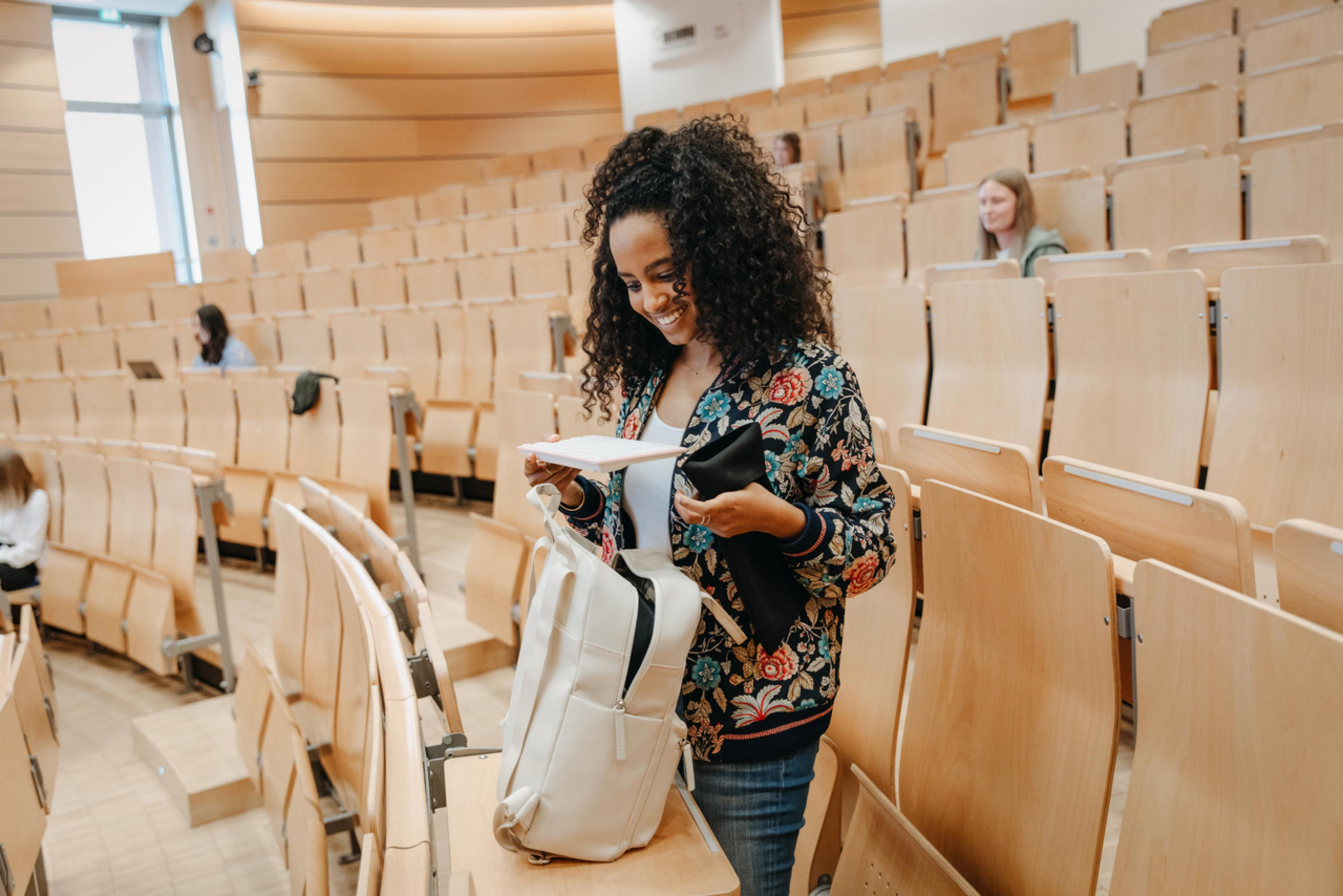 A smiling woman in an auditorium, opening her backpack, with other students in the background.